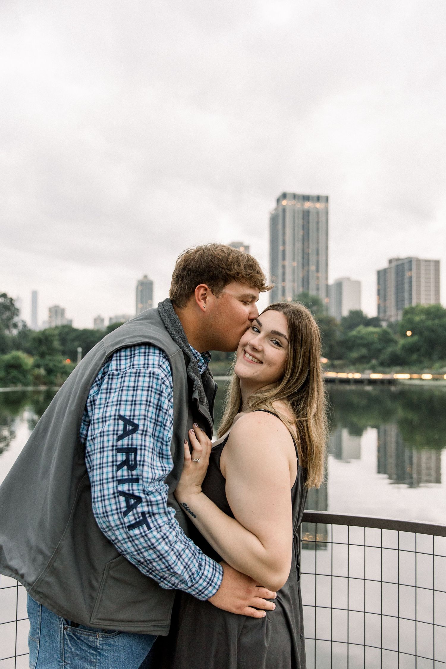 Couple sharing a romantic kiss on a bridge with a city skyline and lake reflection in the background.