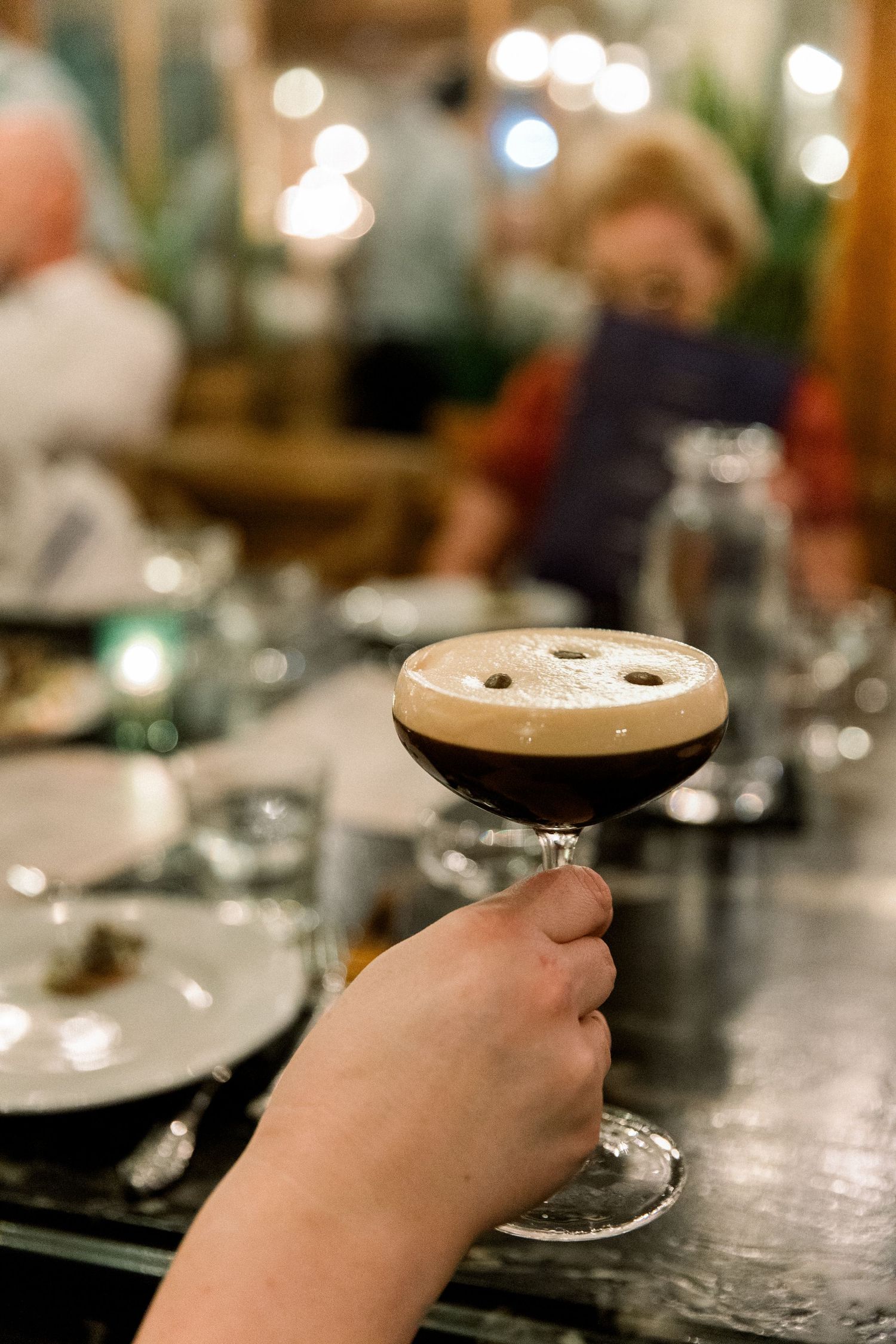 Hand holding espresso martini cocktail with foam top in elegant restaurant setting with bokeh lights.