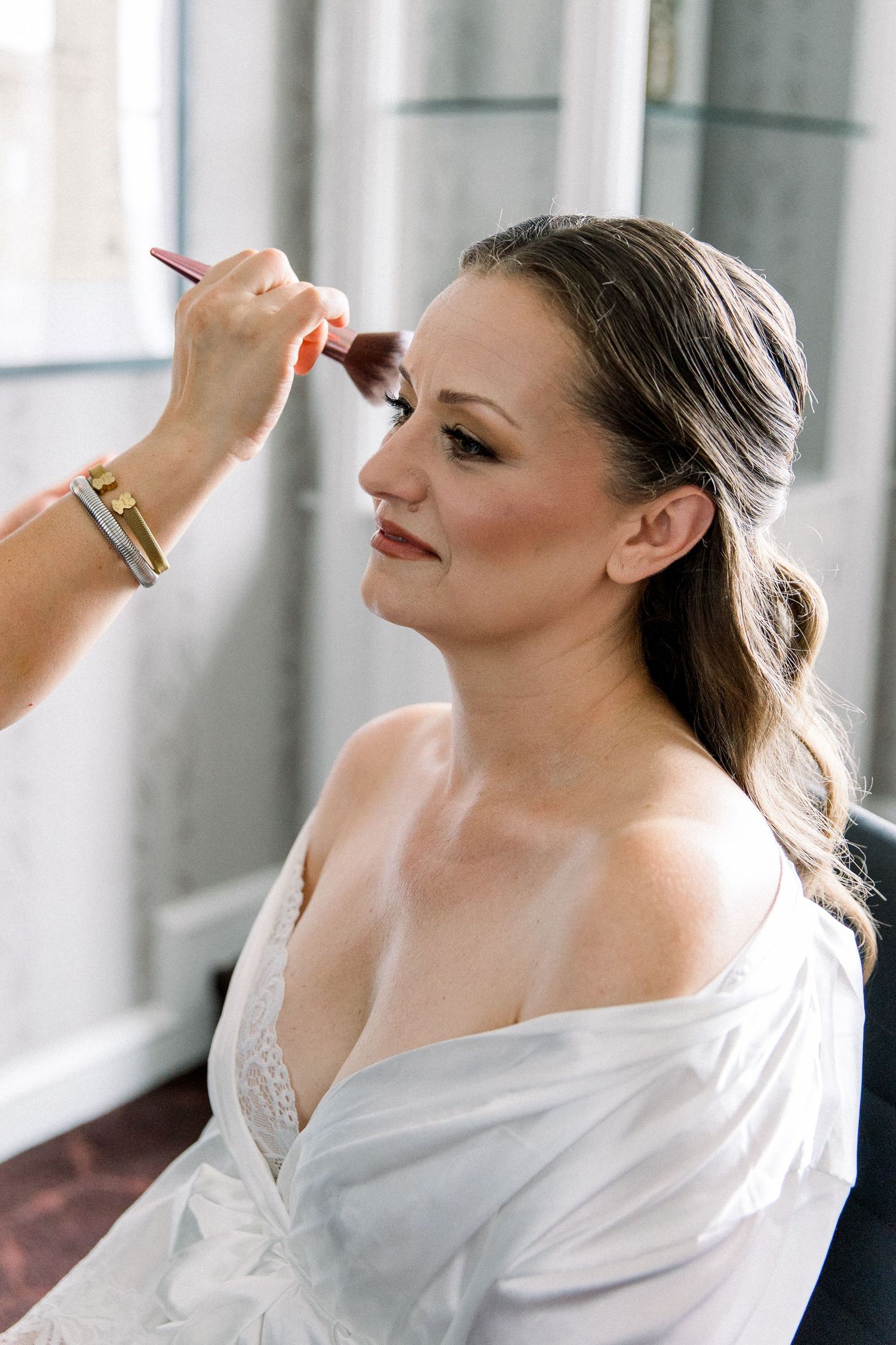 Bride getting makeup applied with a brush while wearing a white robe, preparing for her wedding day.