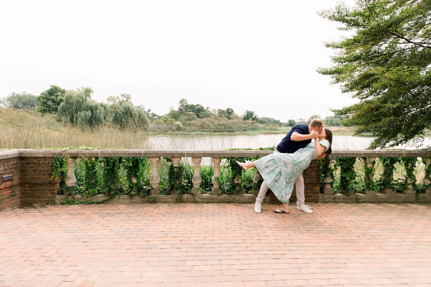 Couple sharing a romantic dip kiss on a brick terrace with stone balustrade and serene lake backdrop.