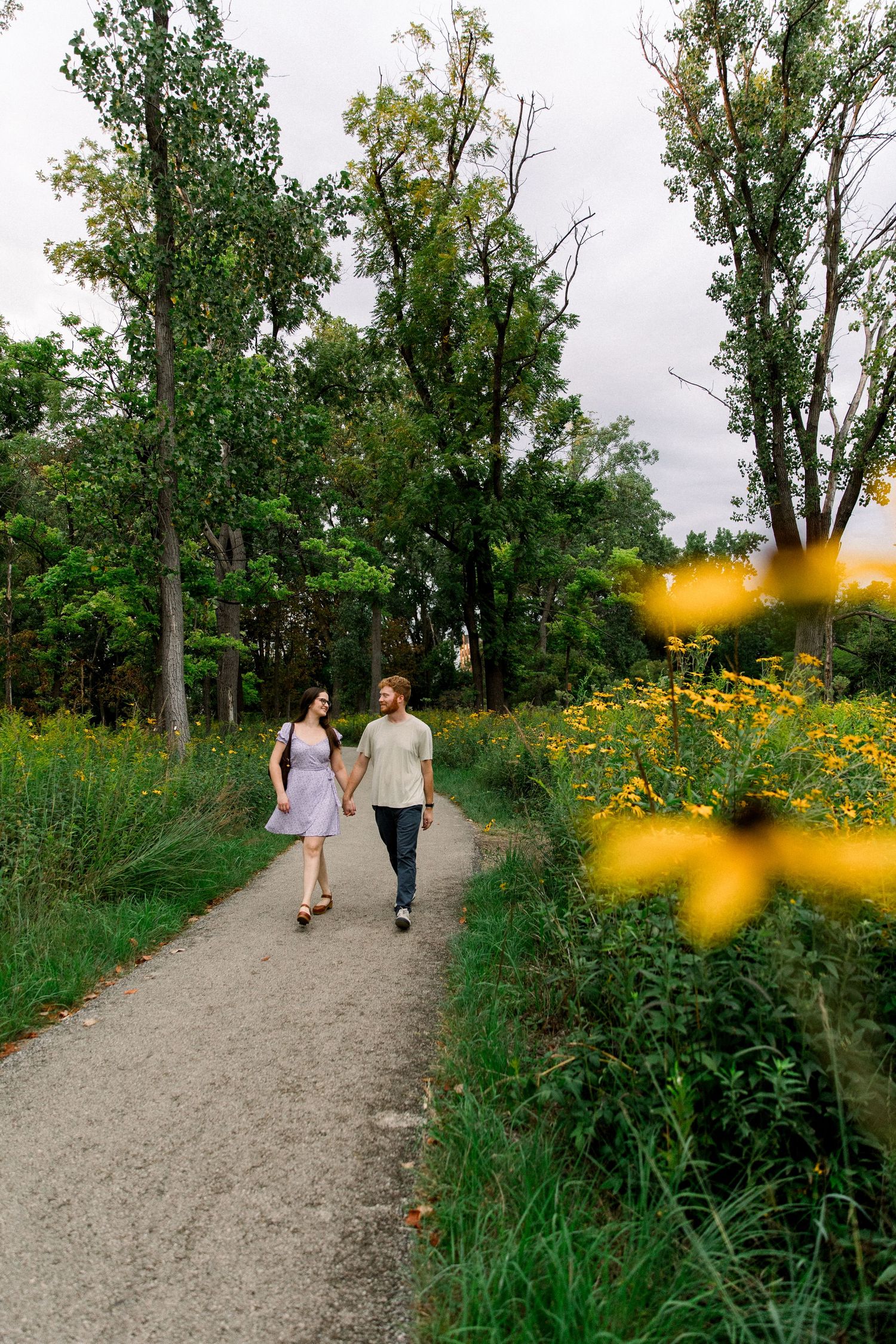 Couple holding hands walking on a scenic path surrounded by yellow wildflowers and lush green trees in a park.