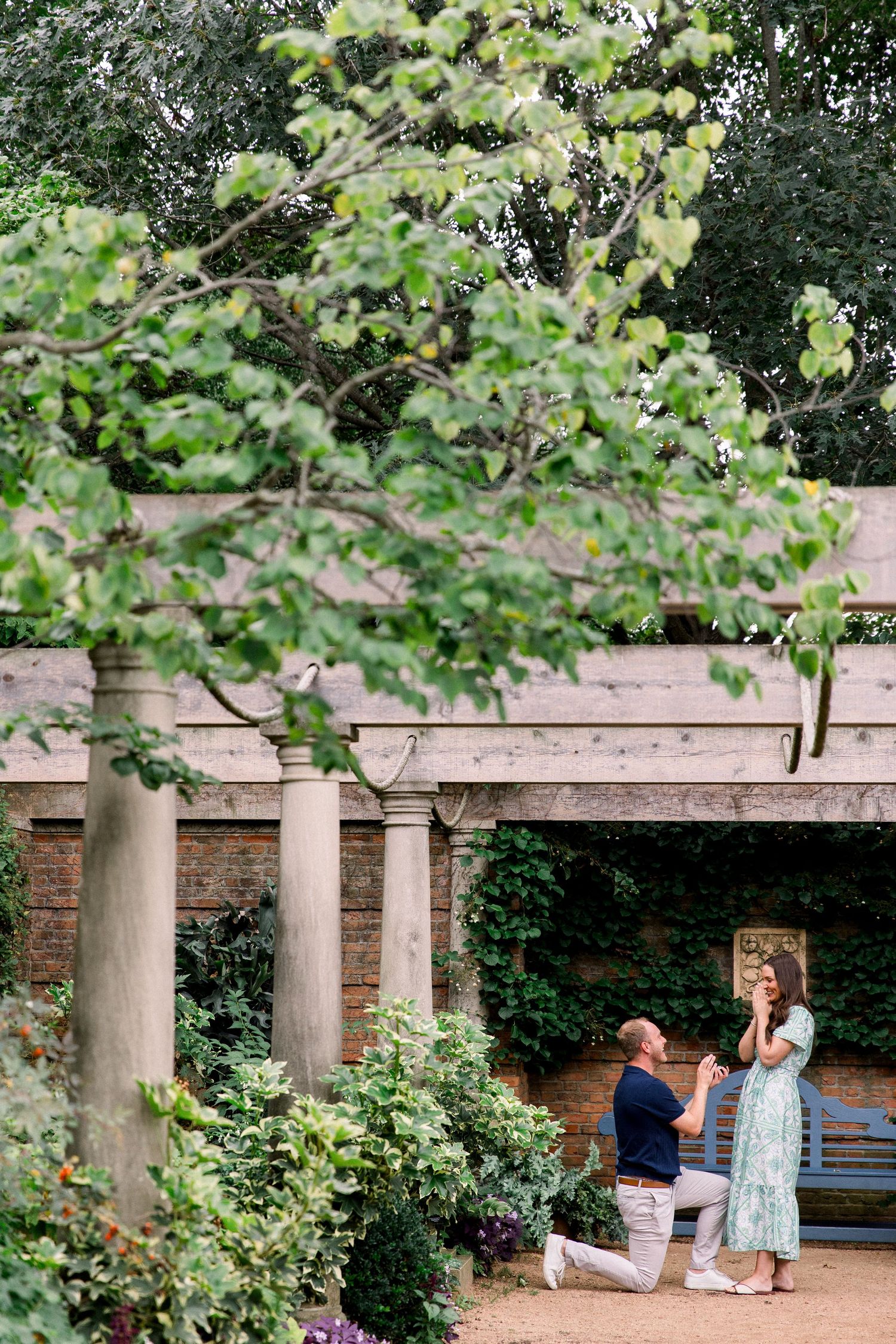 Man proposes to woman on one knee in a lush garden with stone columns and ivy-covered pergola.