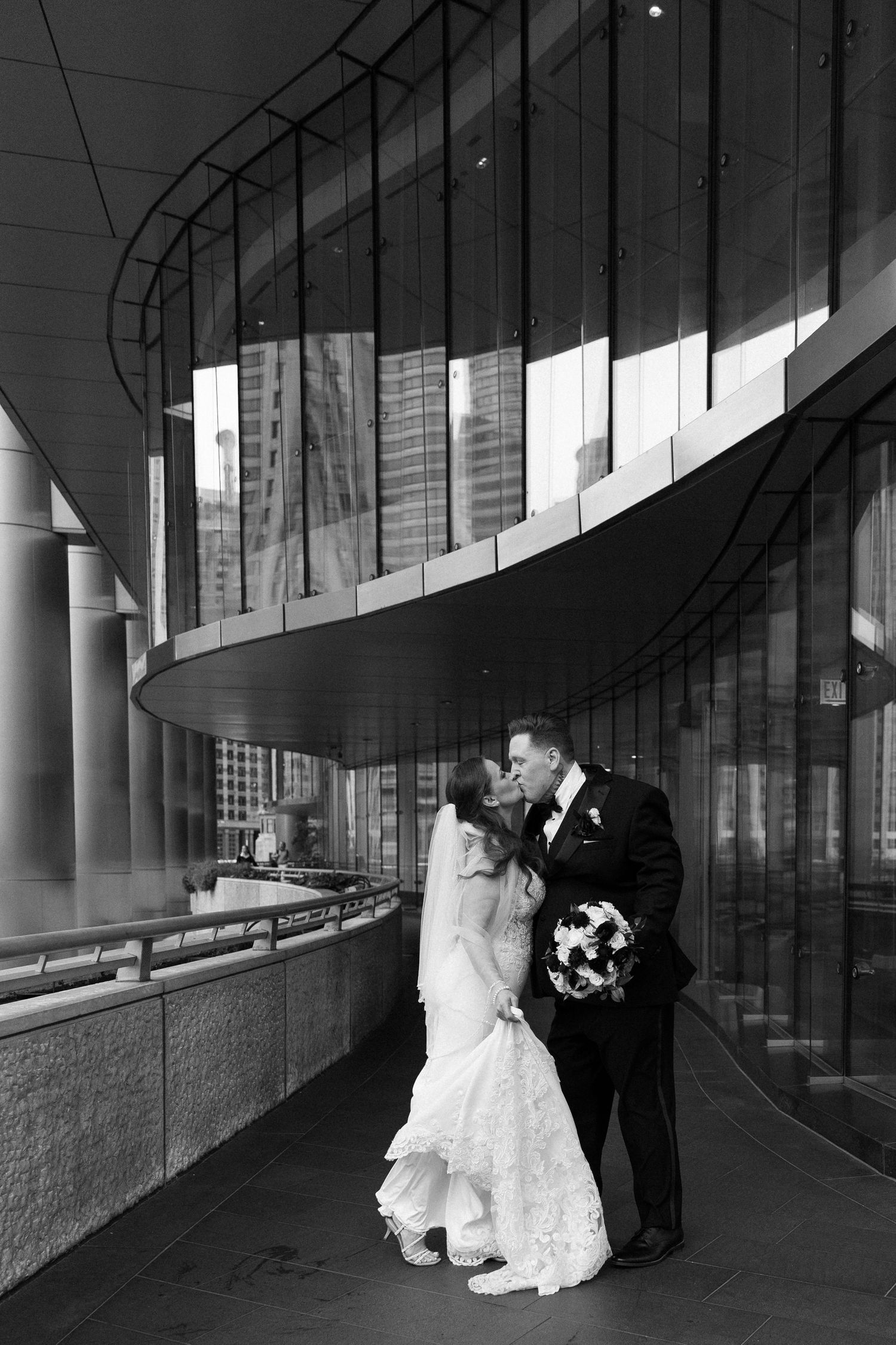 Bride and groom sharing a kiss outside modern glass building in black and white photo.