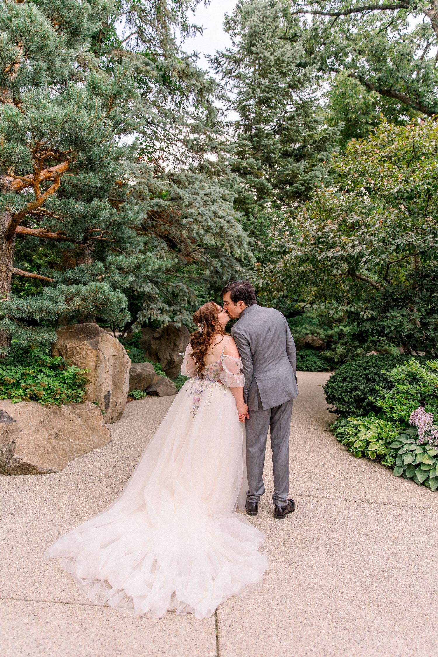 Bride and groom share a kiss on a garden path surrounded by lush green trees and shrubs on their wedding day.