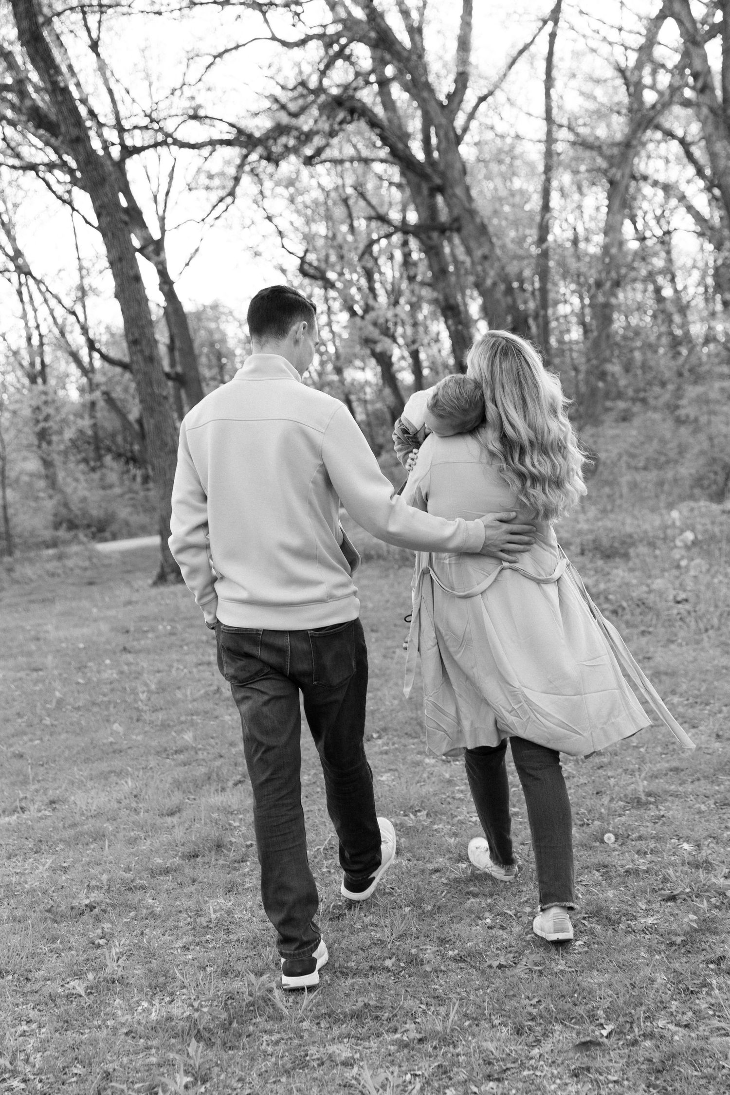 Black and white photo of a family walking through a park, parents embracing child from behind among bare trees.