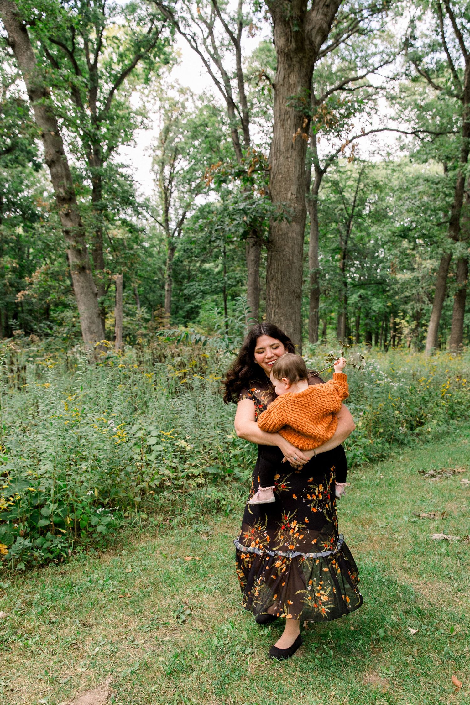 A woman in a floral dress holds a toddler in an orange sweater while walking through a lush green forest.