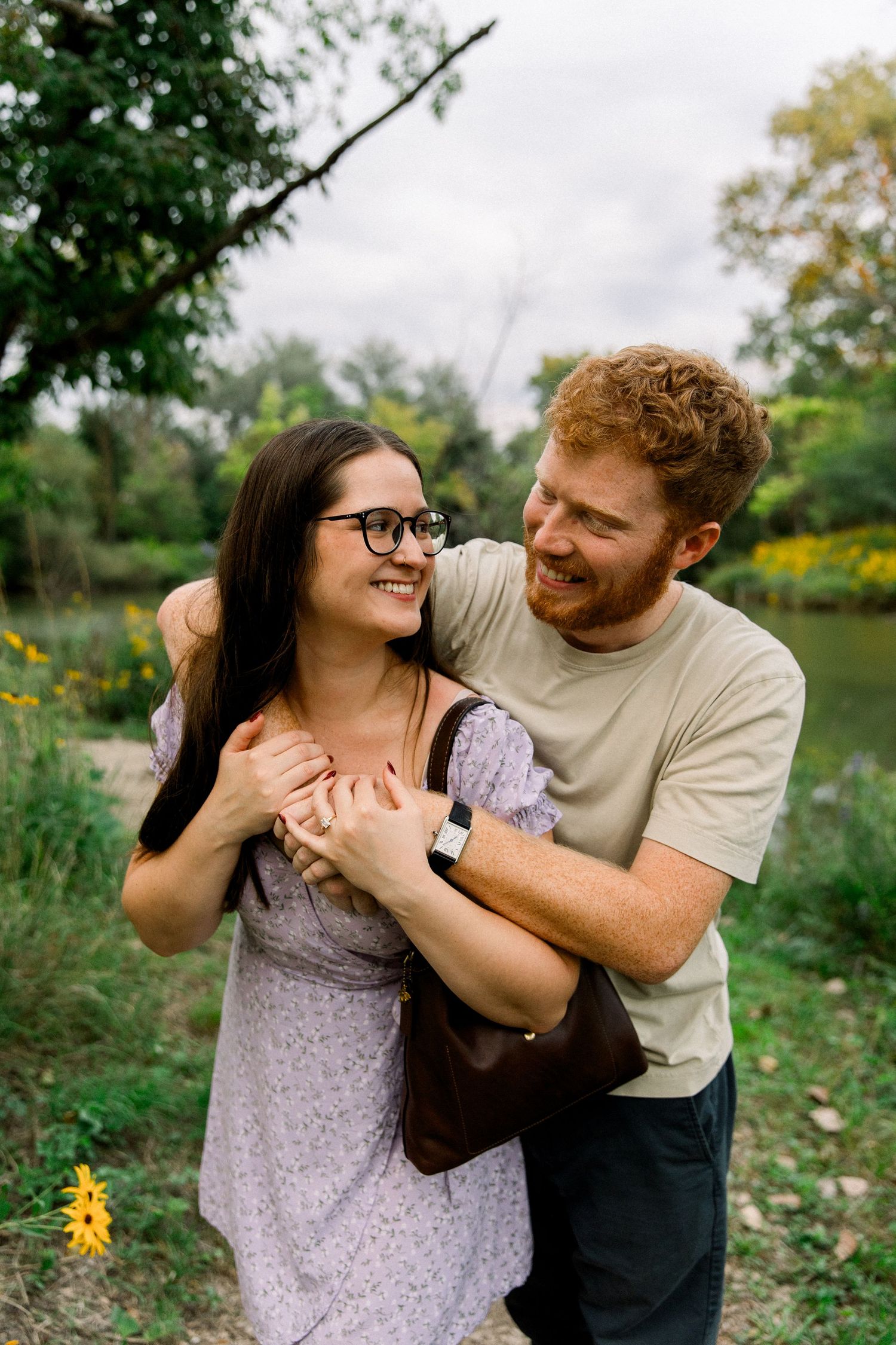 A smiling couple embraces outdoors near a pond, woman in floral dress with glasses, man in beige shirt with red curly hair.