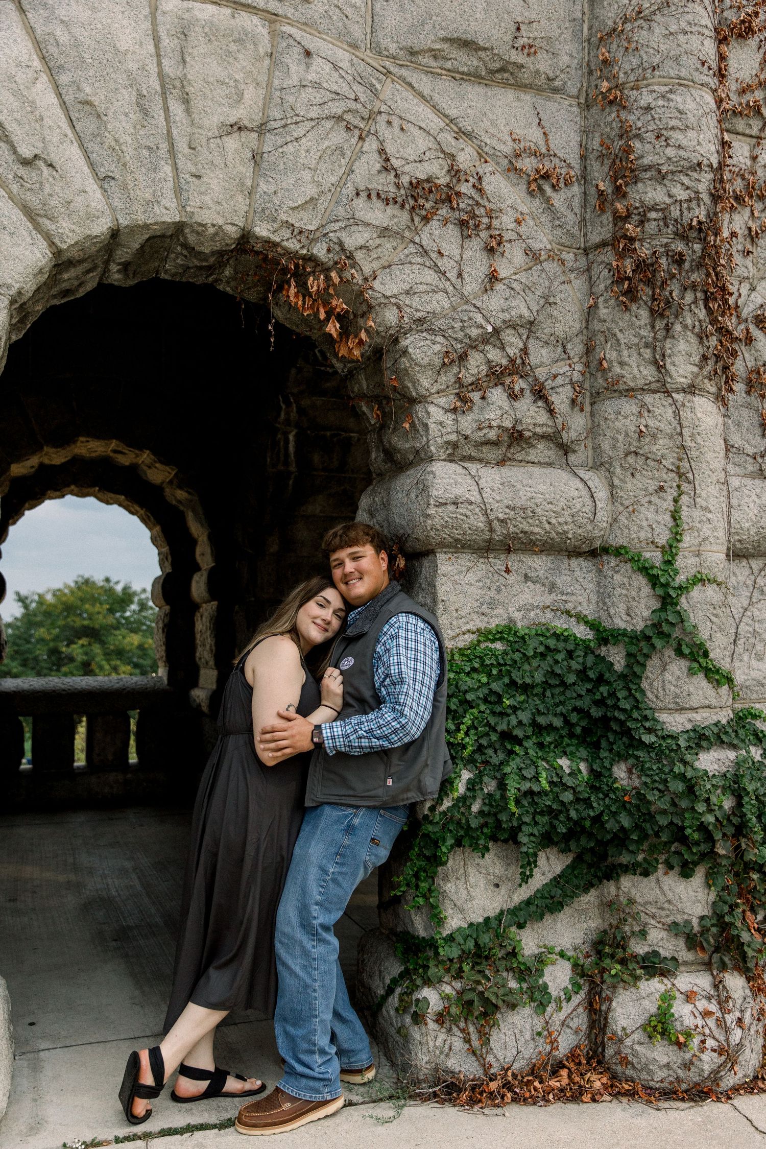Couple snuggling under a stone arch covered in ivy, woman in black dress leans into man in plaid shirt and vest.
