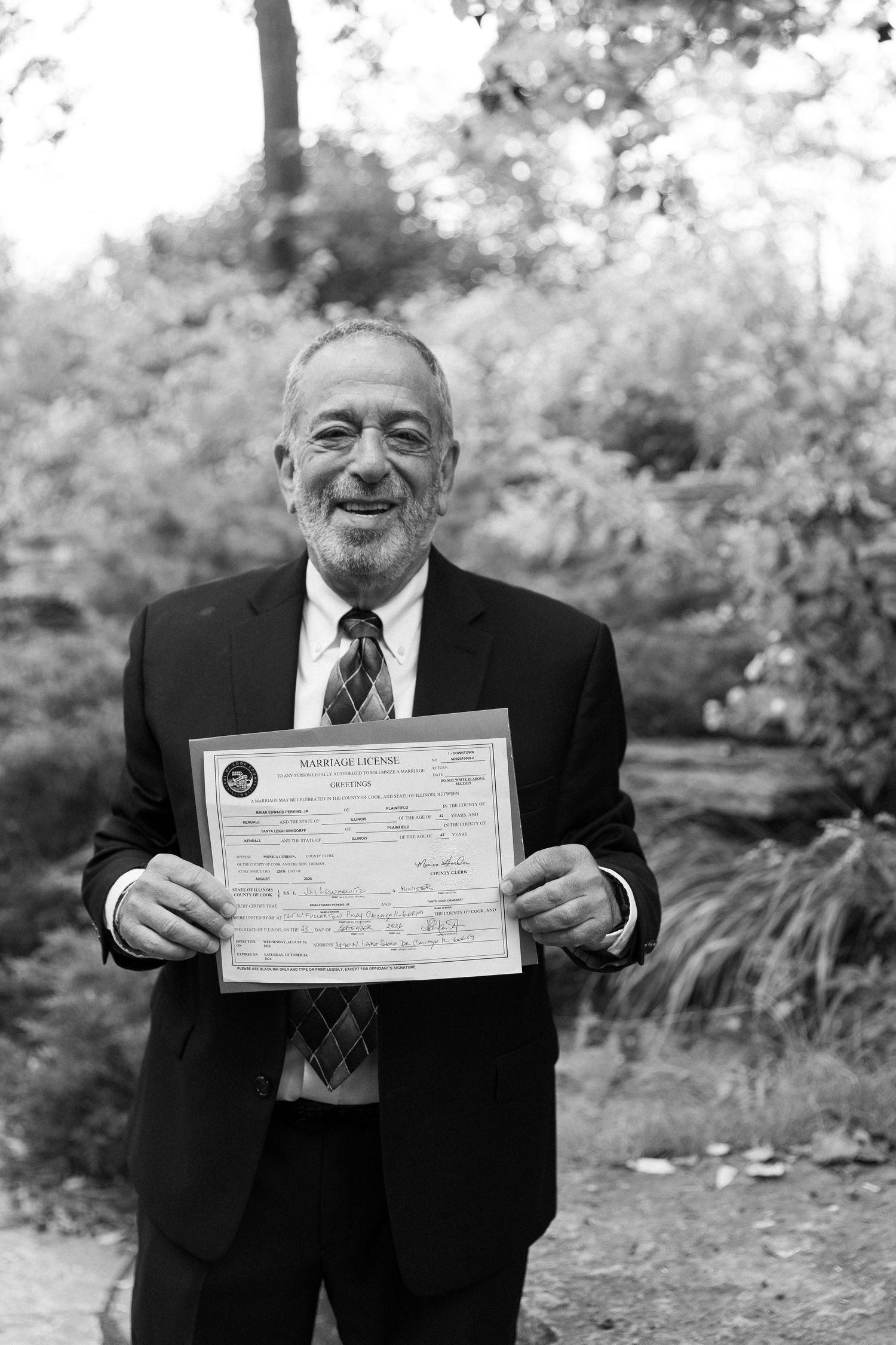 Smiling older gentleman in suit proudly displays an official marriage license certificate outdoors in black and white photo.
