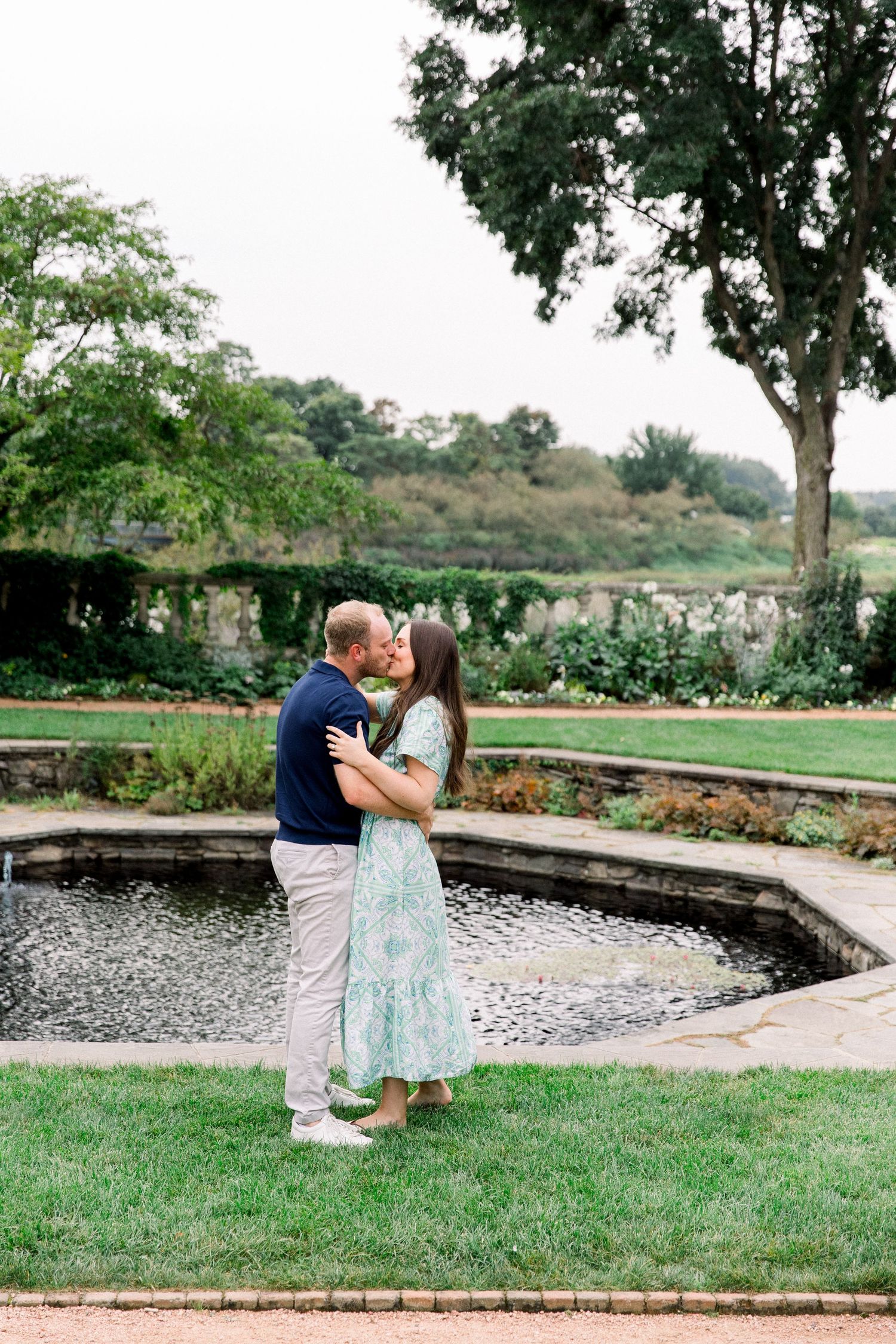 Couple kissing by a serene garden pond with green landscape backdrop.