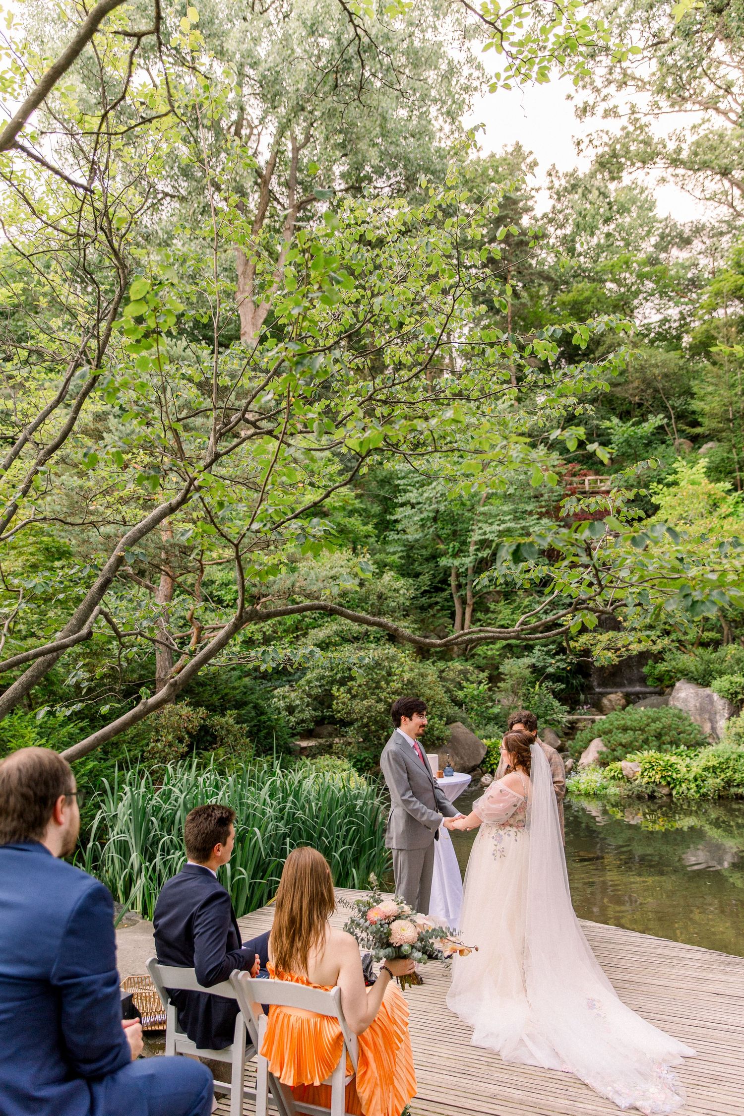 Outdoor wedding ceremony on a wooden deck surrounded by lush greenery and a pond, with guests seated watching the couple exchange vows.