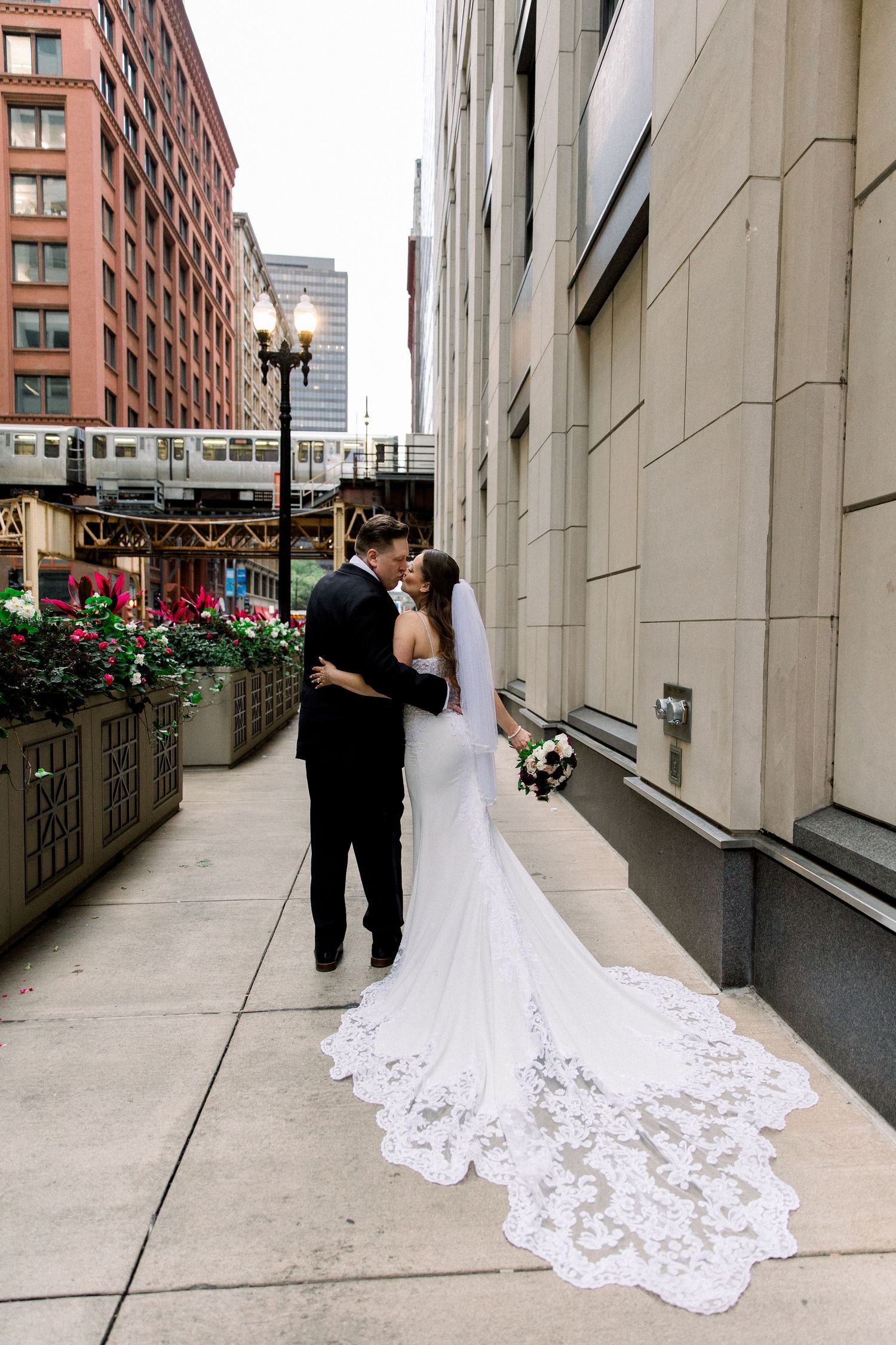 Bride and groom share a romantic kiss on a Chicago city sidewalk, her lace cathedral veil trailing elegantly behind her.