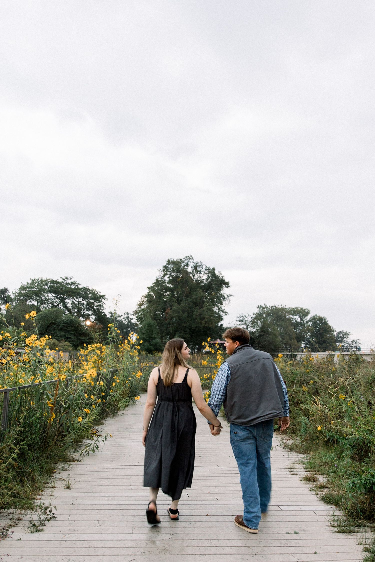 Couple holding hands walking on a wooden boardwalk path through yellow wildflowers on a cloudy day.