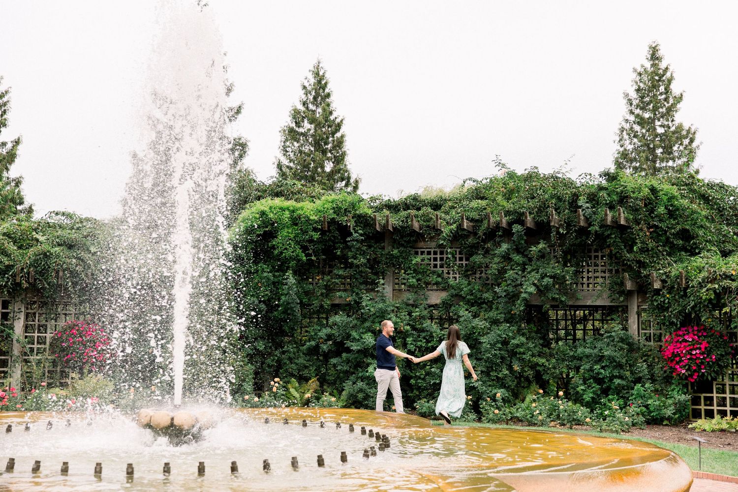 Couple dancing by a garden fountain surrounded by lush greenery and colorful flowers on a cloudy day.