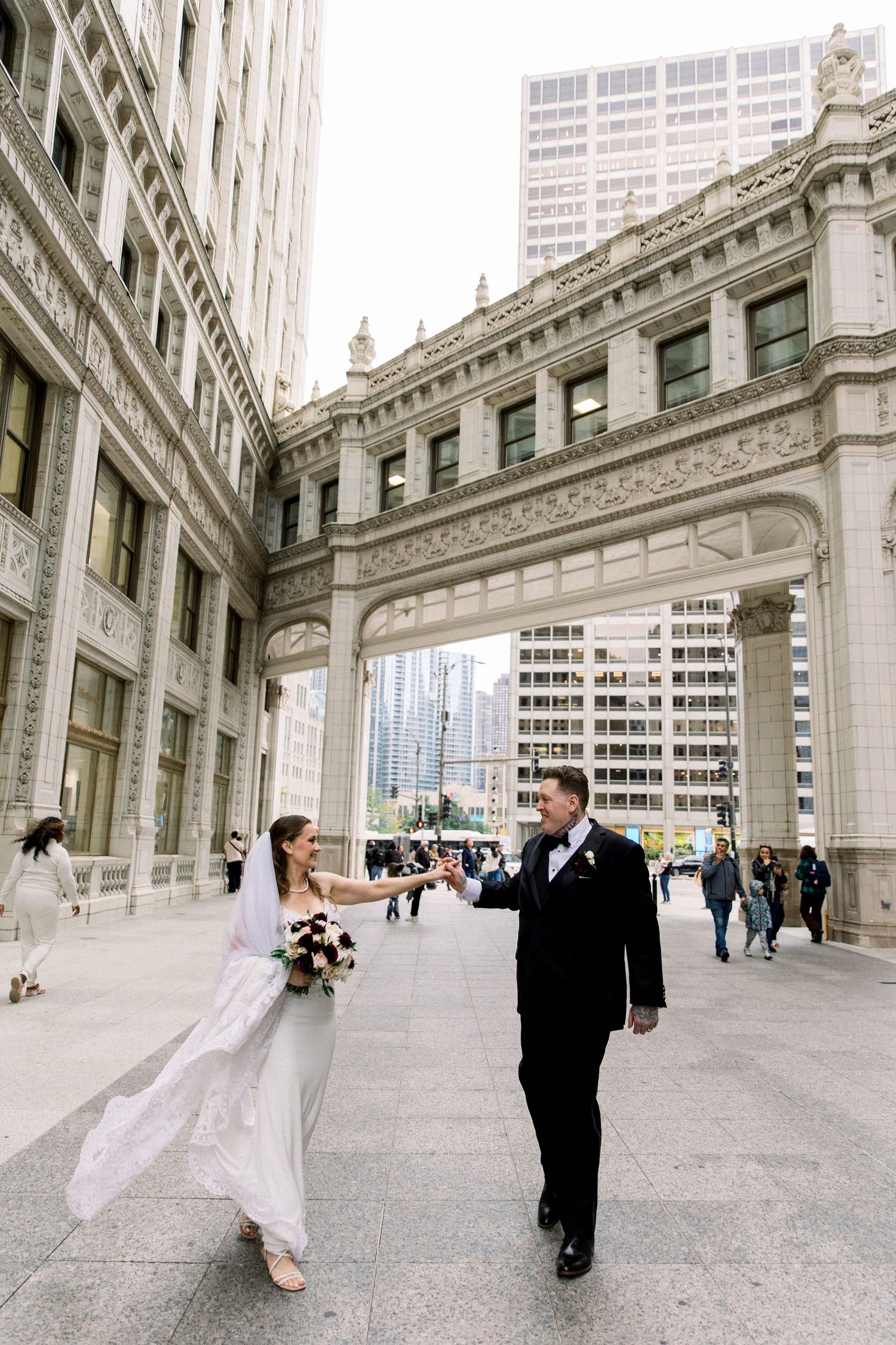 Bride and groom holding hands walking near ornate Chicago bridge architecture on wedding day.