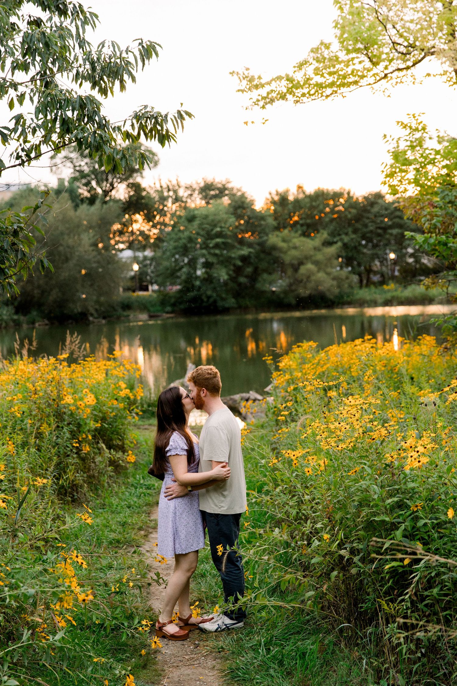 Couple sharing a kiss among vibrant yellow wildflowers beside a serene lake at sunset in a green park.