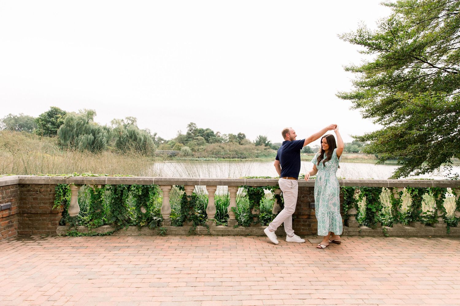 Couple dancing on a brick terrace overlooking a serene lake surrounded by lush greenery.