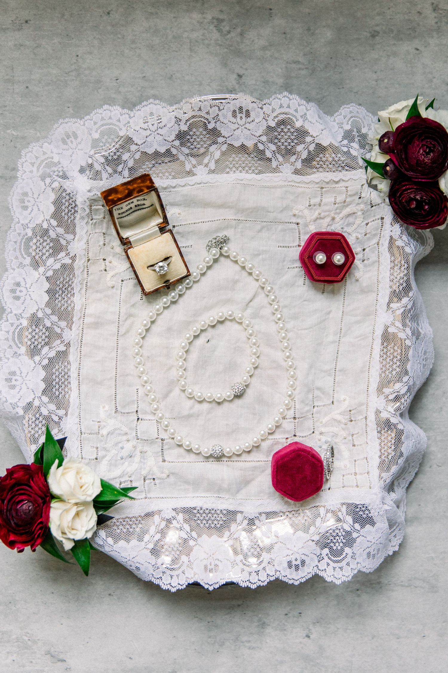 Bridal flat lay with pearl necklace, diamond ring, red earrings, and roses on lace fabric.