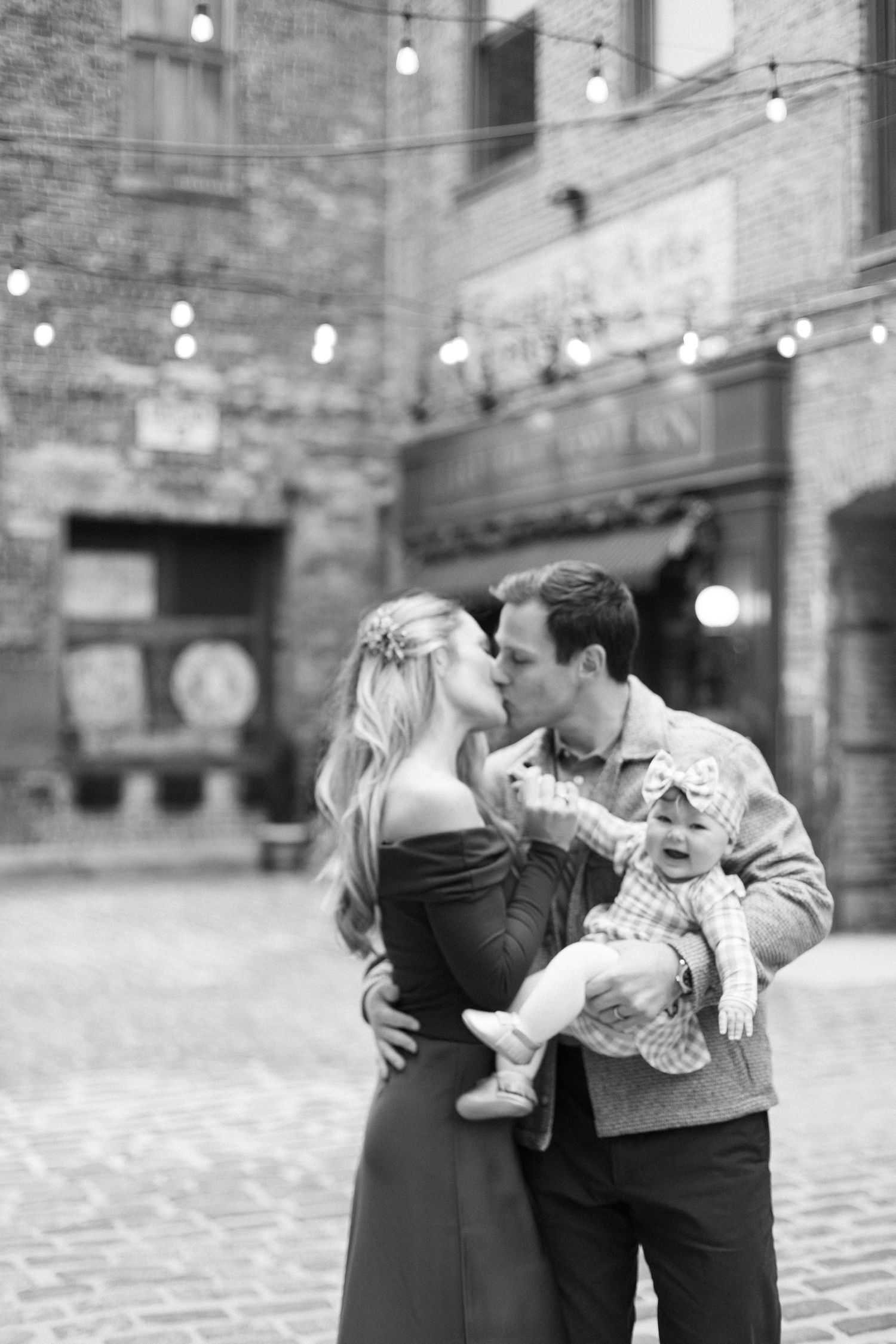 Black and white photo of a couple kissing while holding their baby in a charming brick courtyard with string lights.