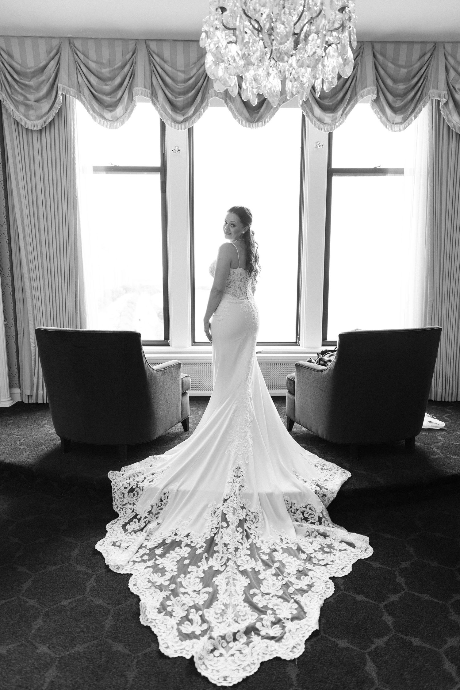 Bride in elegant lace train wedding dress standing by window in black and white photo.