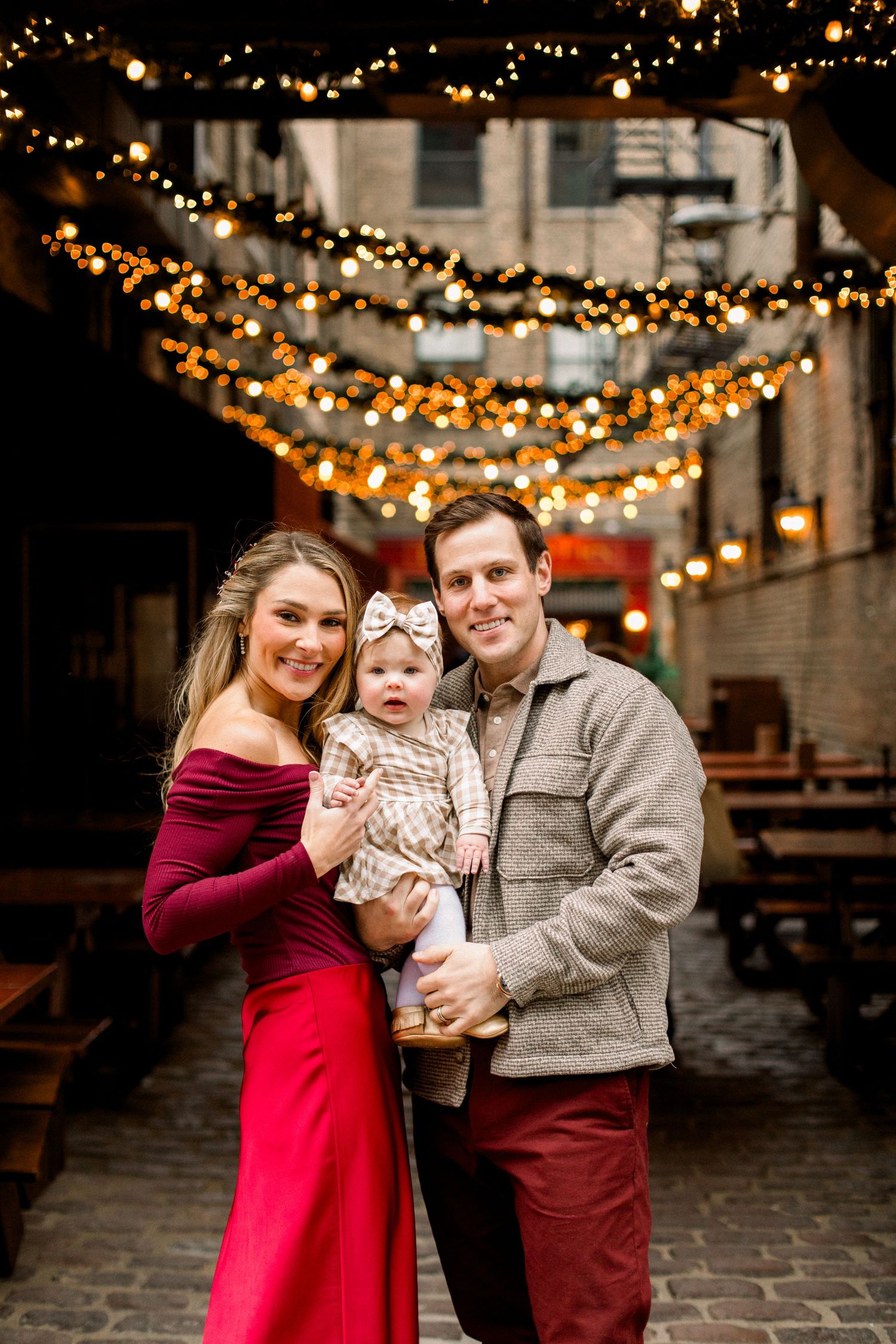 A smiling family of three poses under warm string lights in a festive outdoor alley, parents holding their baby girl.