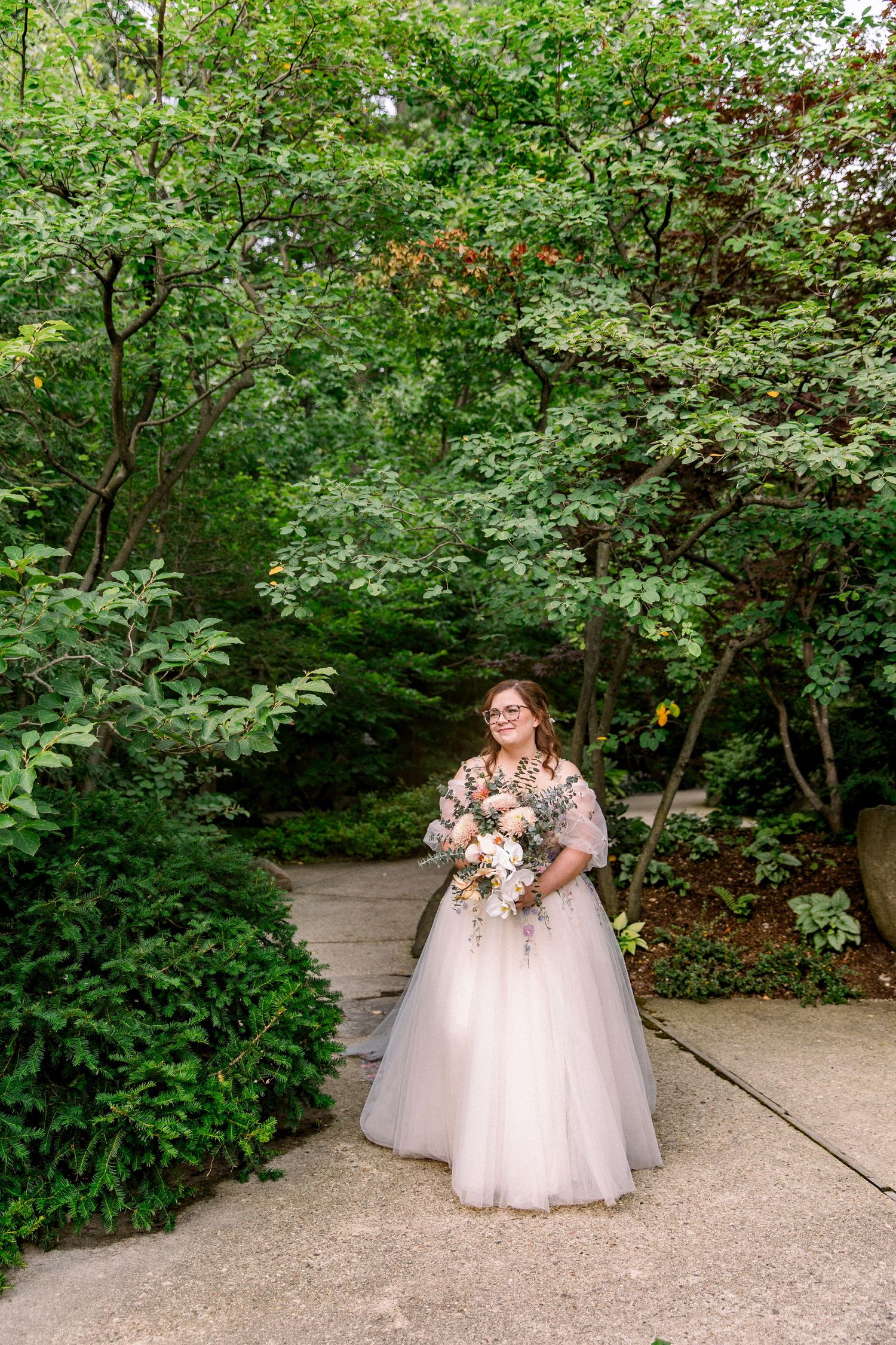 Bride in blush tulle ball gown holding floral bouquet stands on garden path surrounded by lush green trees.