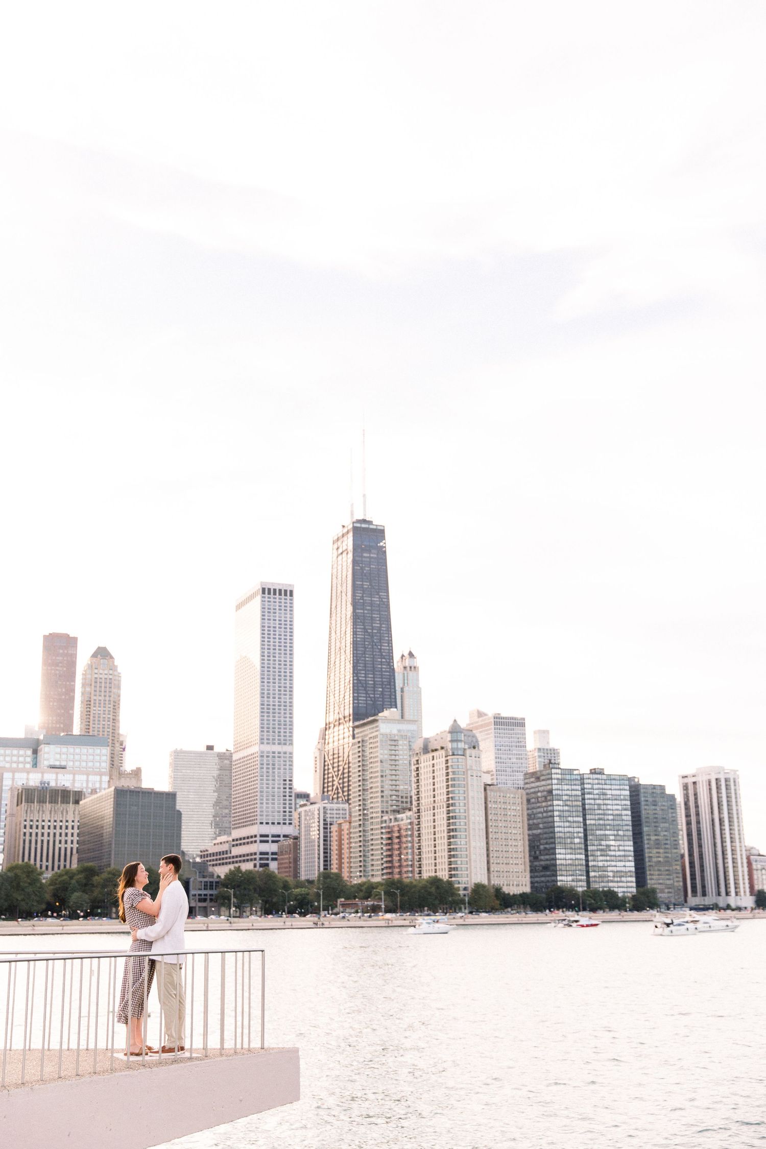 Romantic couple embracing on lakefront pier with stunning Chicago skyline and John Hancock Tower in the background.