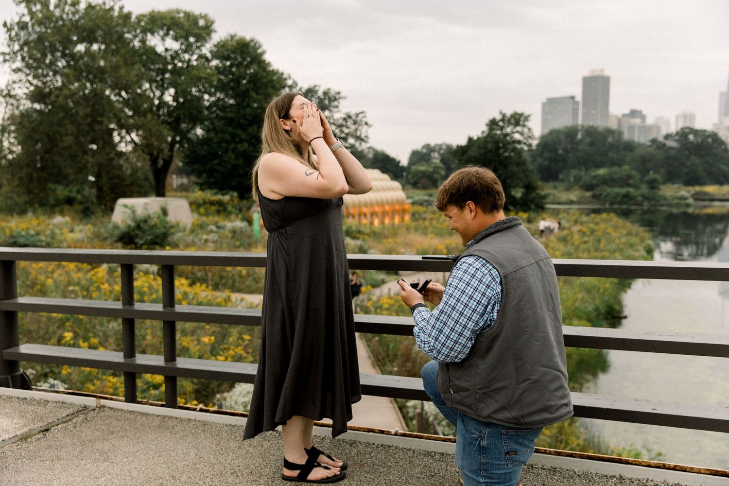 Man proposes on a bridge as woman covers her face in surprise, with a city skyline and pond in the background.