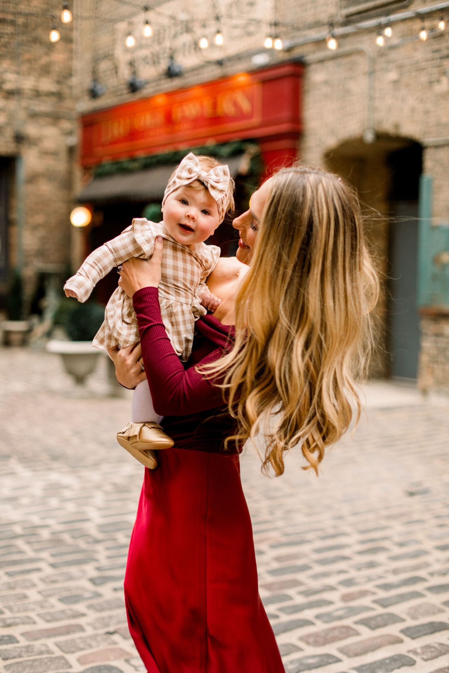 Woman in red dress holds smiling baby in plaid outfit with bow headband outdoors on cobblestone street.