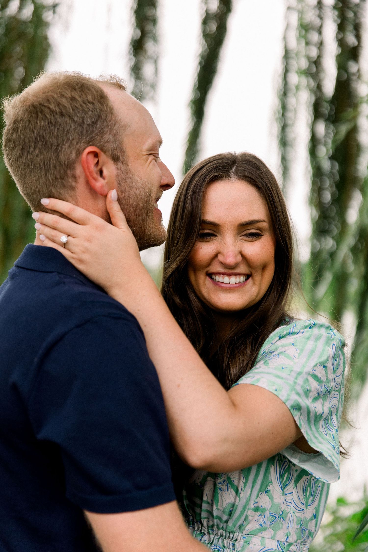Smiling woman in green dress holds man's face tenderly during romantic engagement photo session outdoors.