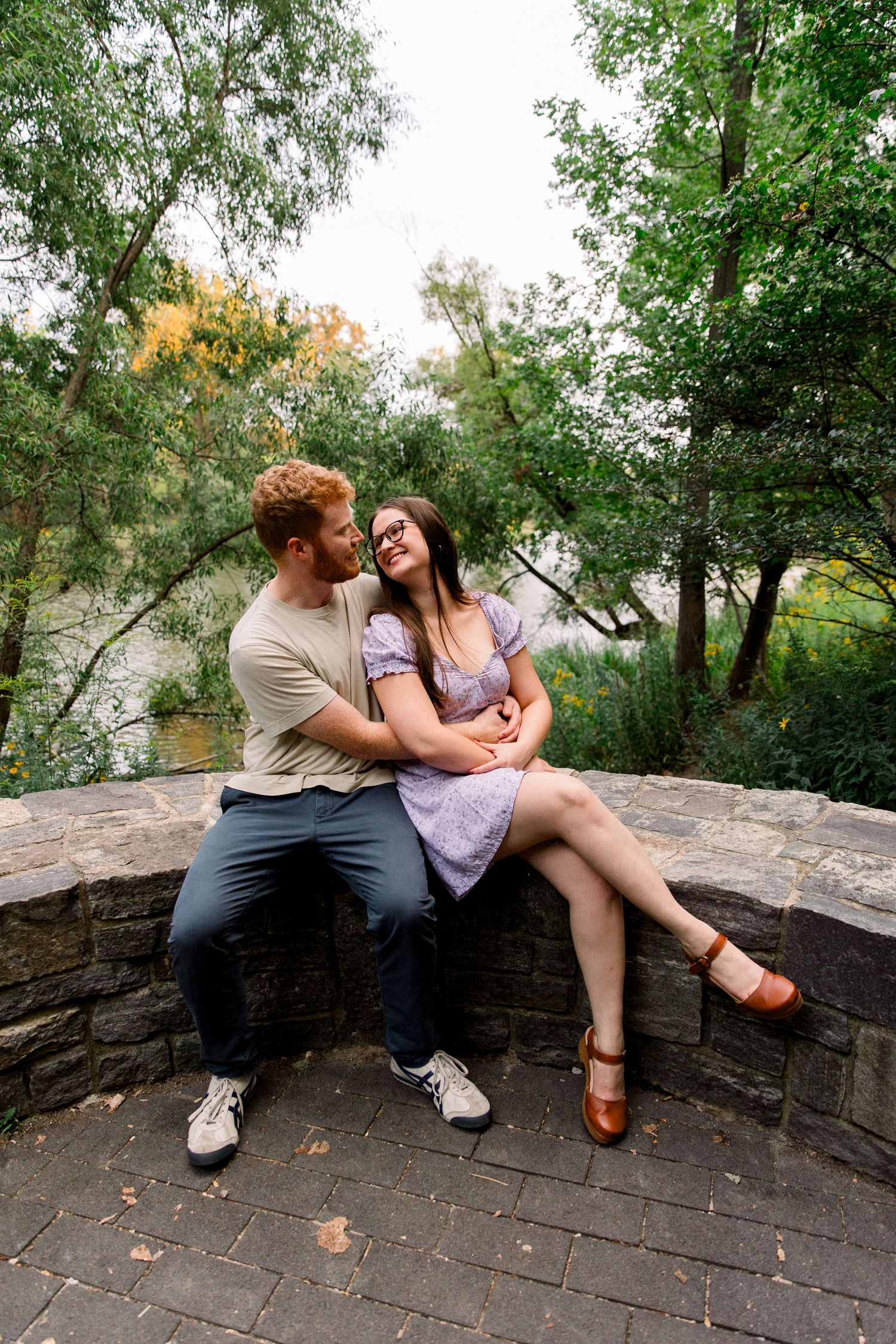 A couple sits closely on a stone wall surrounded by lush green trees, sharing a tender moment outdoors.