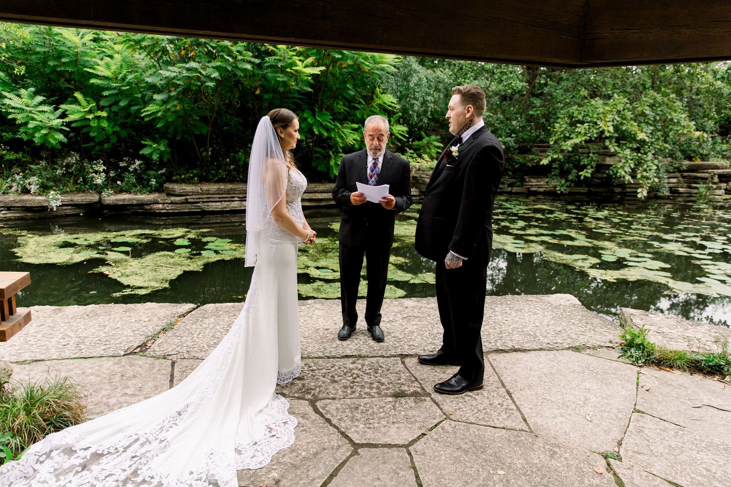 Bride and groom exchange vows with officiant beside a serene lily pond in a lush garden wedding ceremony.
