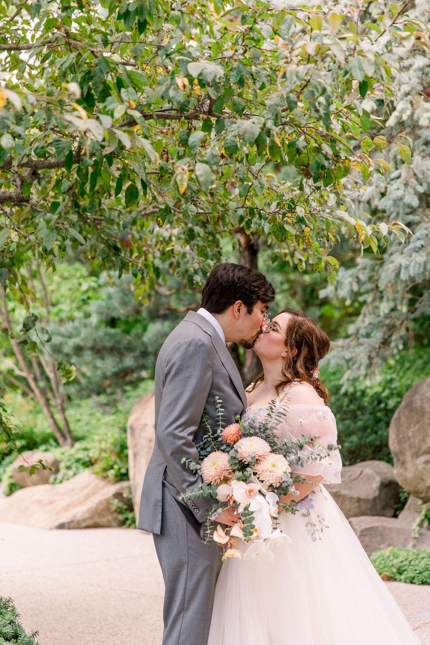 Bride and groom share a romantic kiss under a leafy tree, bride holding a soft peach and white floral bouquet.