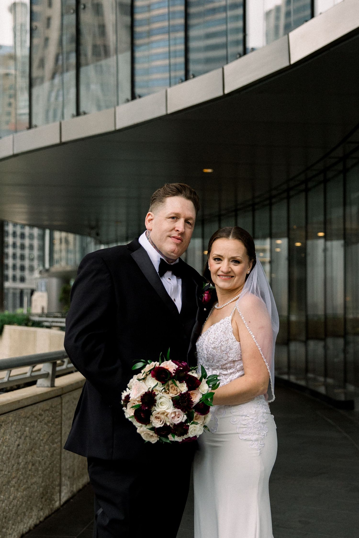 Newlyweds pose together on urban terrace, bride holding bouquet of burgundy and cream roses in white gown.