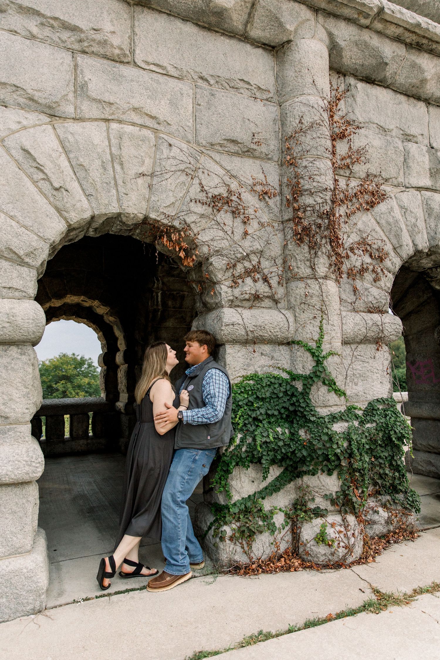 Couple embracing against a stone arch wall with ivy, sharing a romantic moment outdoors.