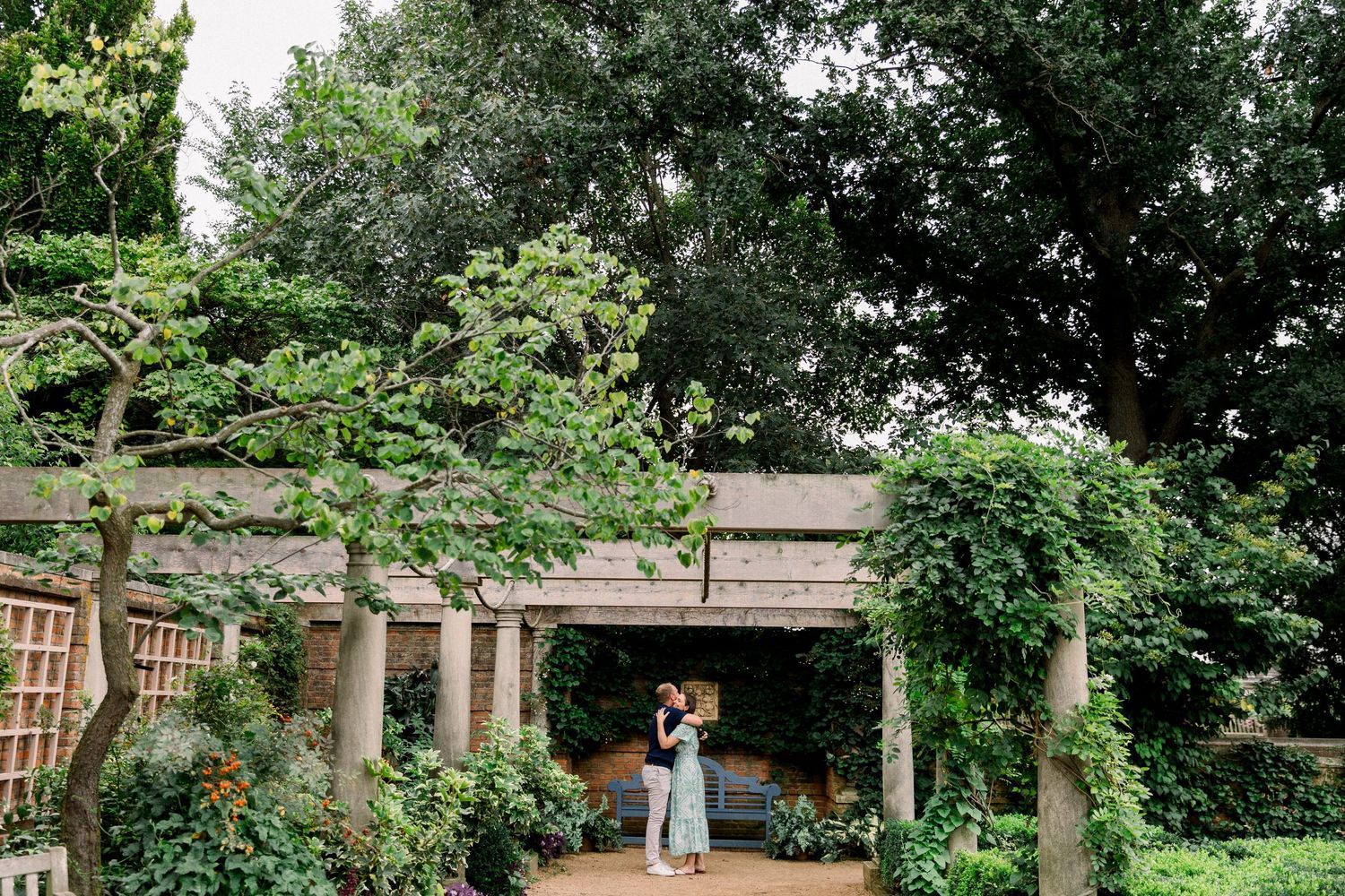 Couple embracing under a rustic pergola surrounded by lush greenery in a romantic garden setting.
