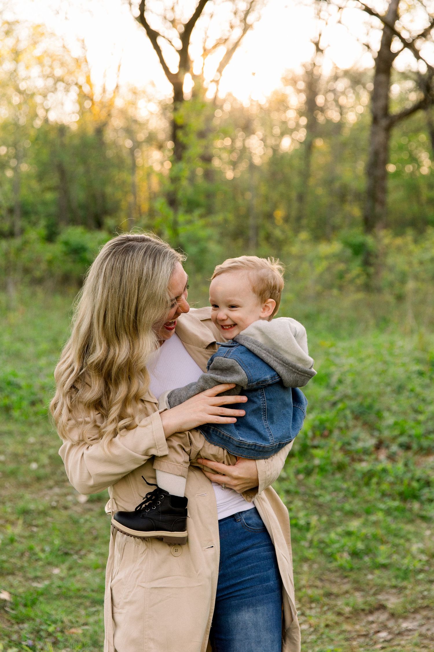 A smiling woman in a tan coat holds a laughing toddler boy outdoors in a sunlit green forest setting.