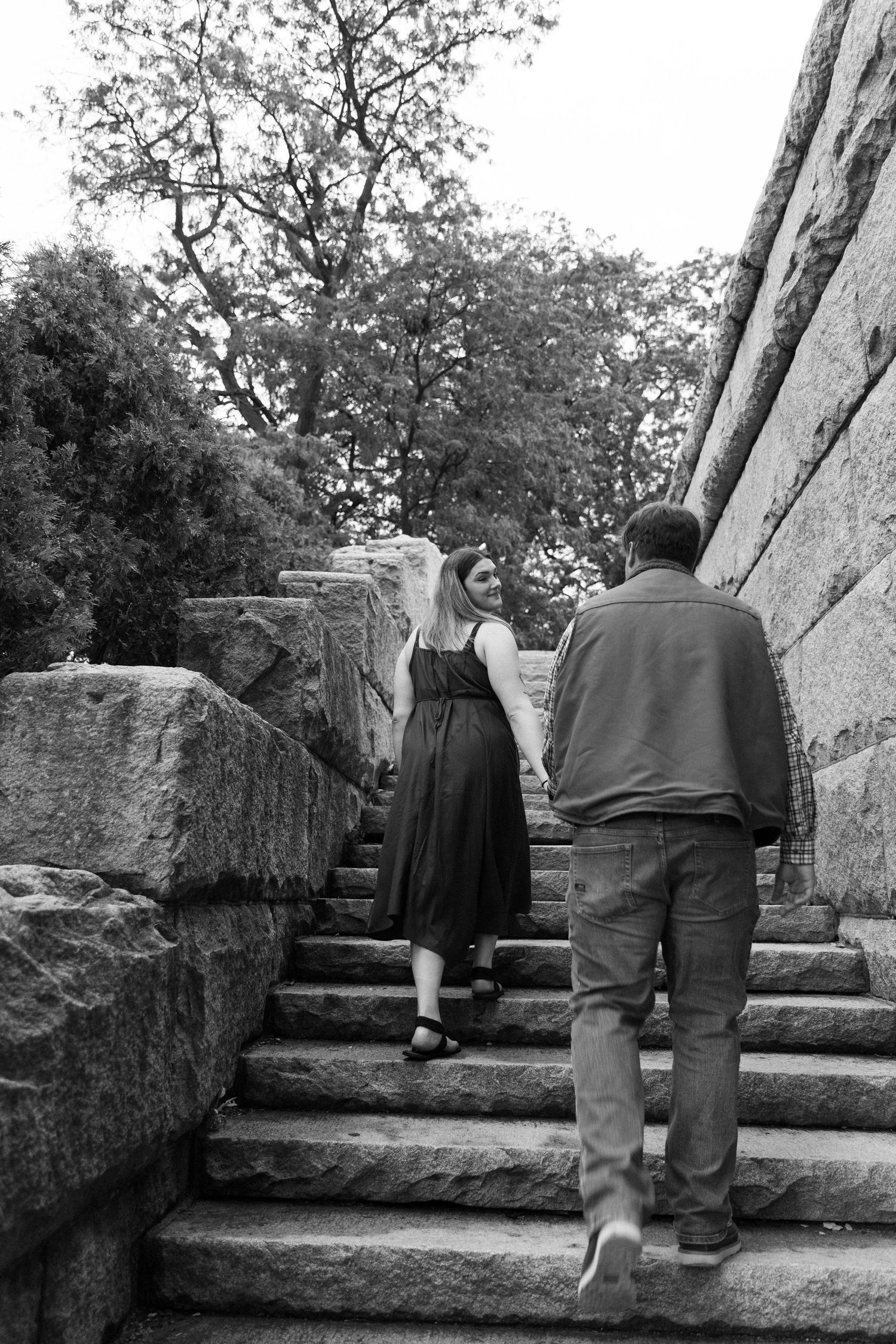 Couple climbing stone stairs outdoors, woman in black dress looks back at man in blue jacket, black and white photo.