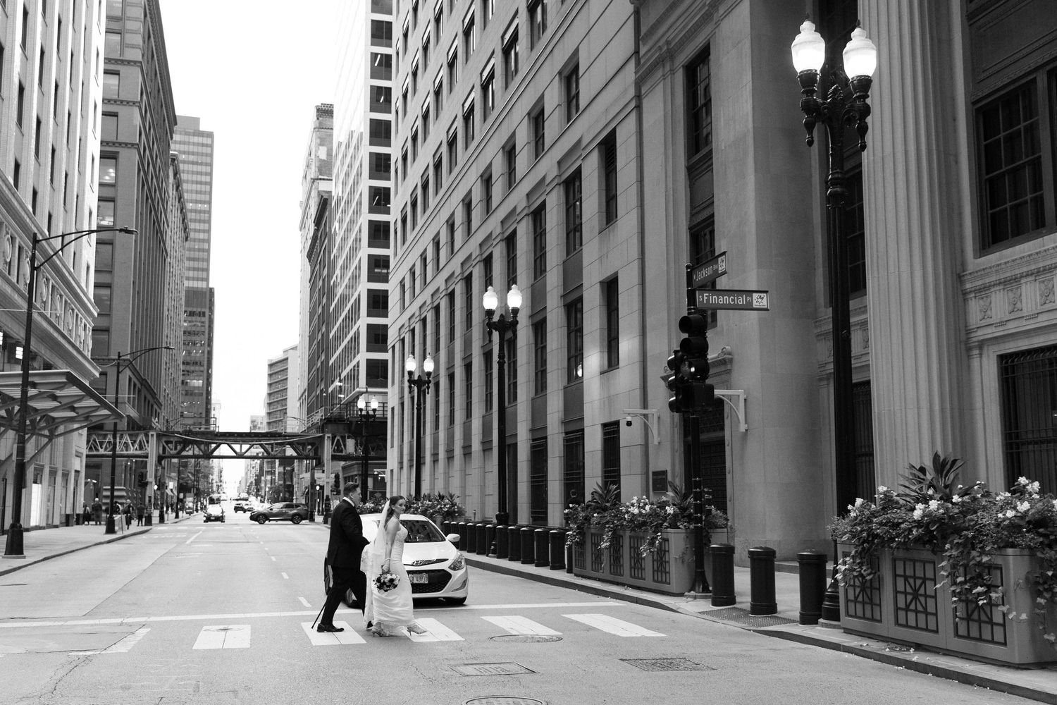 Black and white photo of a bride and groom crossing a city street amid tall buildings and a parked car.