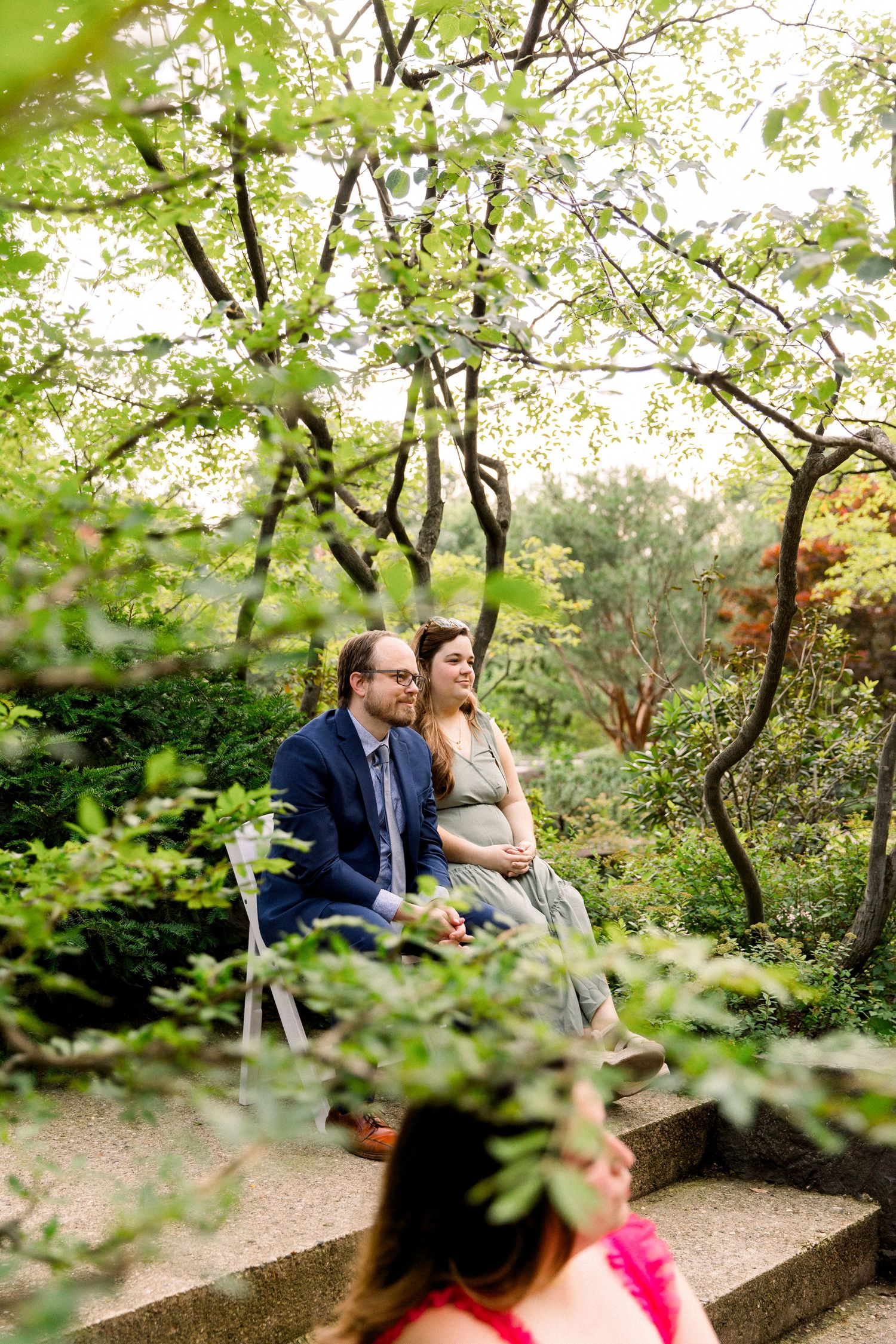 Couple sits together on stone steps surrounded by blooming green trees during an outdoor garden wedding ceremony.