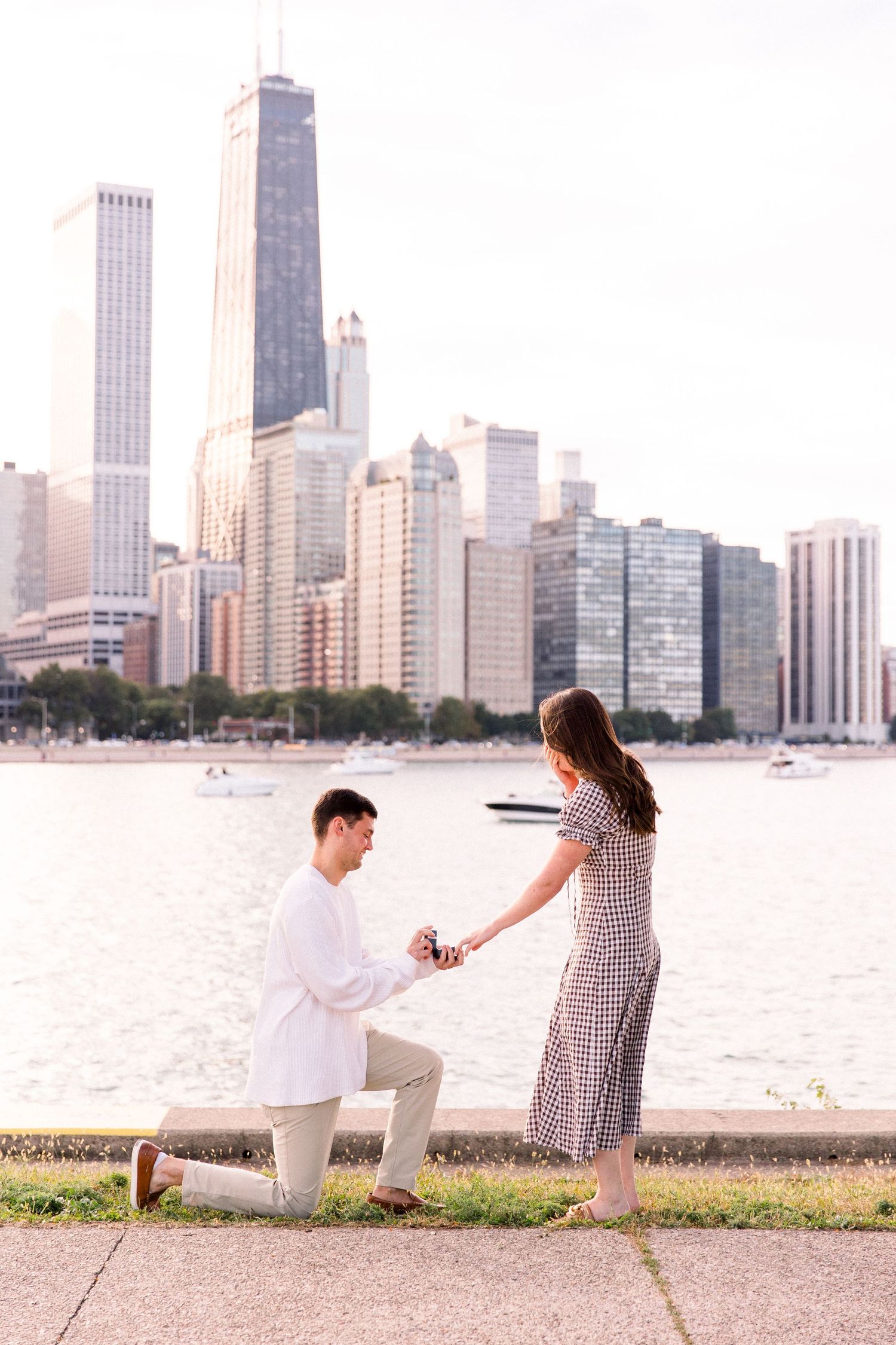 Man proposes on one knee to woman by Chicago skyline waterfront on sunny day.