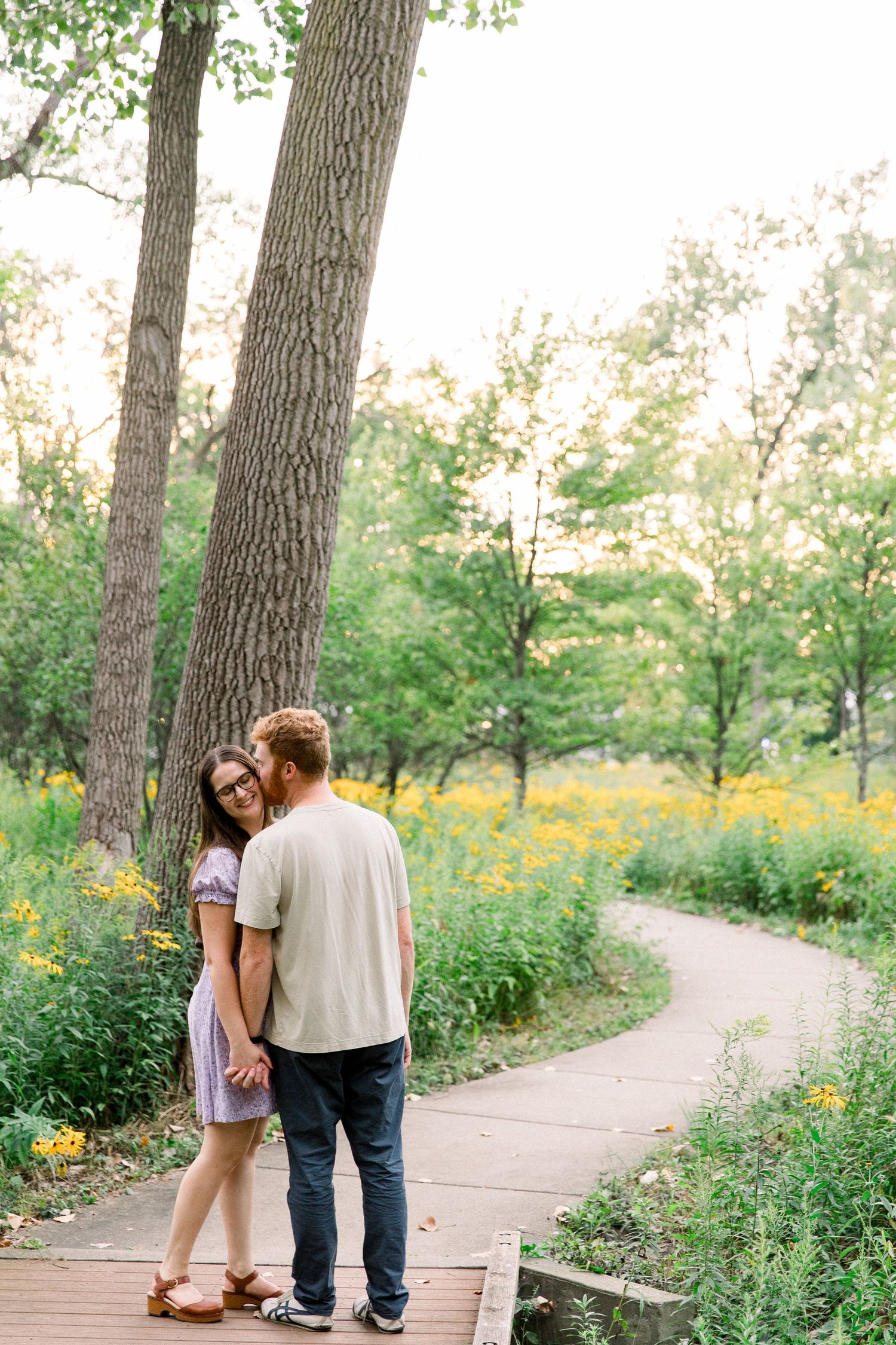Romantic couple holding hands on a winding wooden boardwalk path through a lush park with yellow flowers.