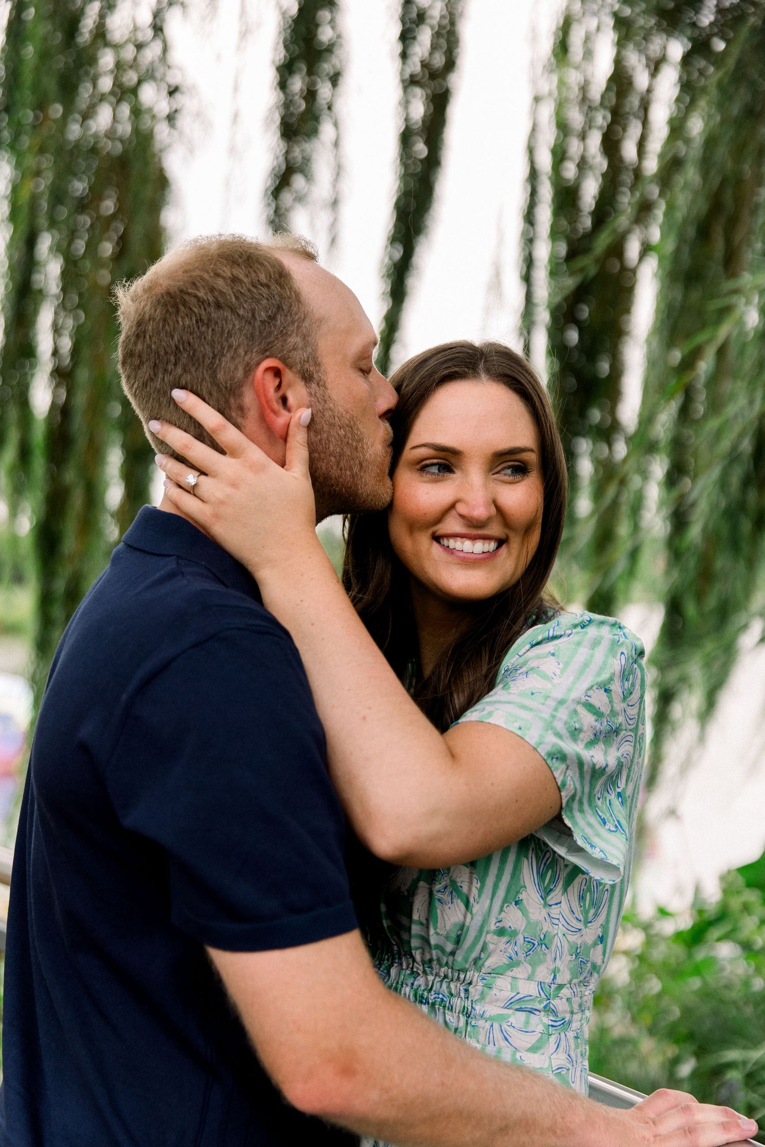 A man kisses a smiling woman on the cheek outdoors, she wears a green dress and an engagement ring.