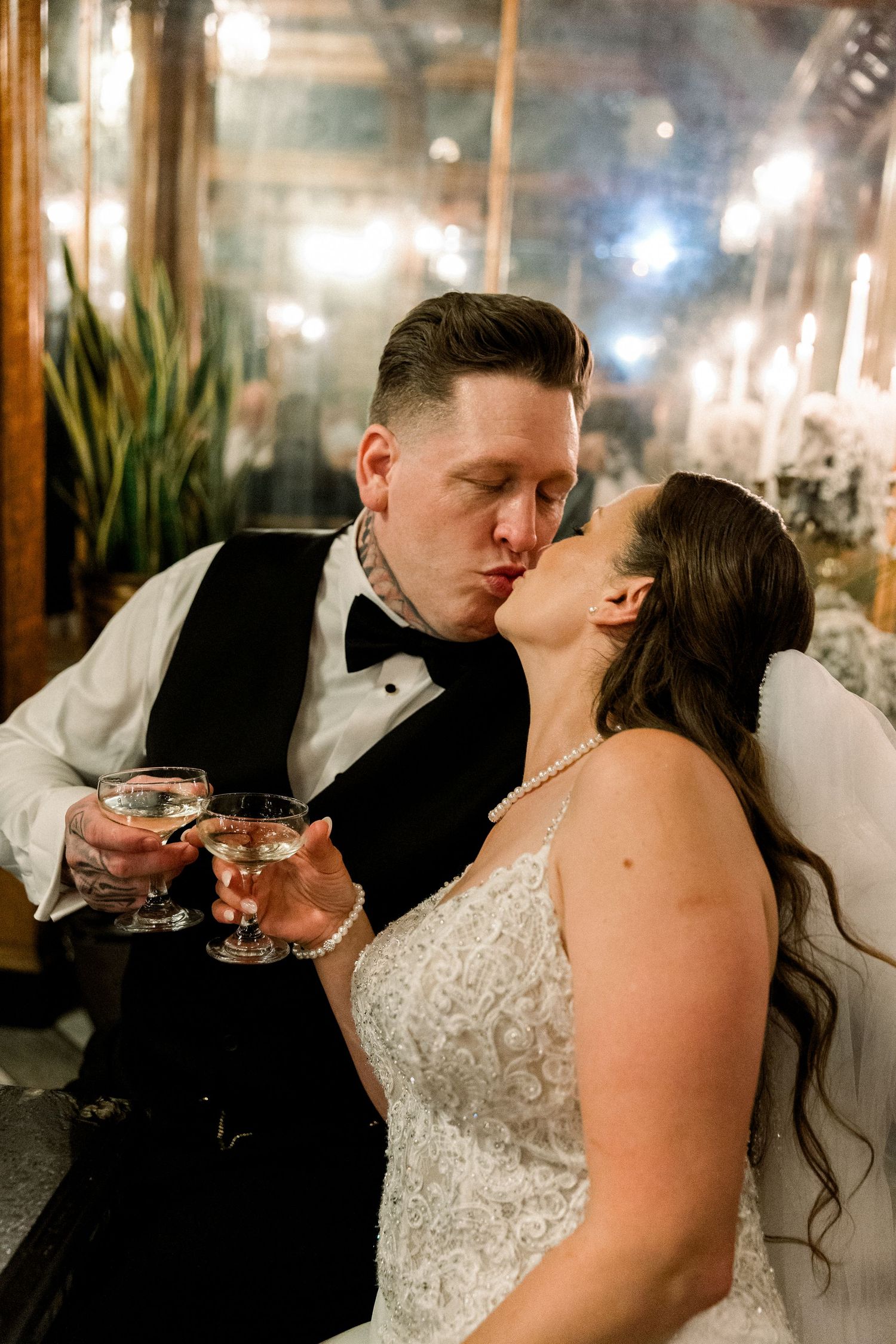 Newlyweds share a kiss while holding champagne glasses at their elegant candlelit wedding reception.