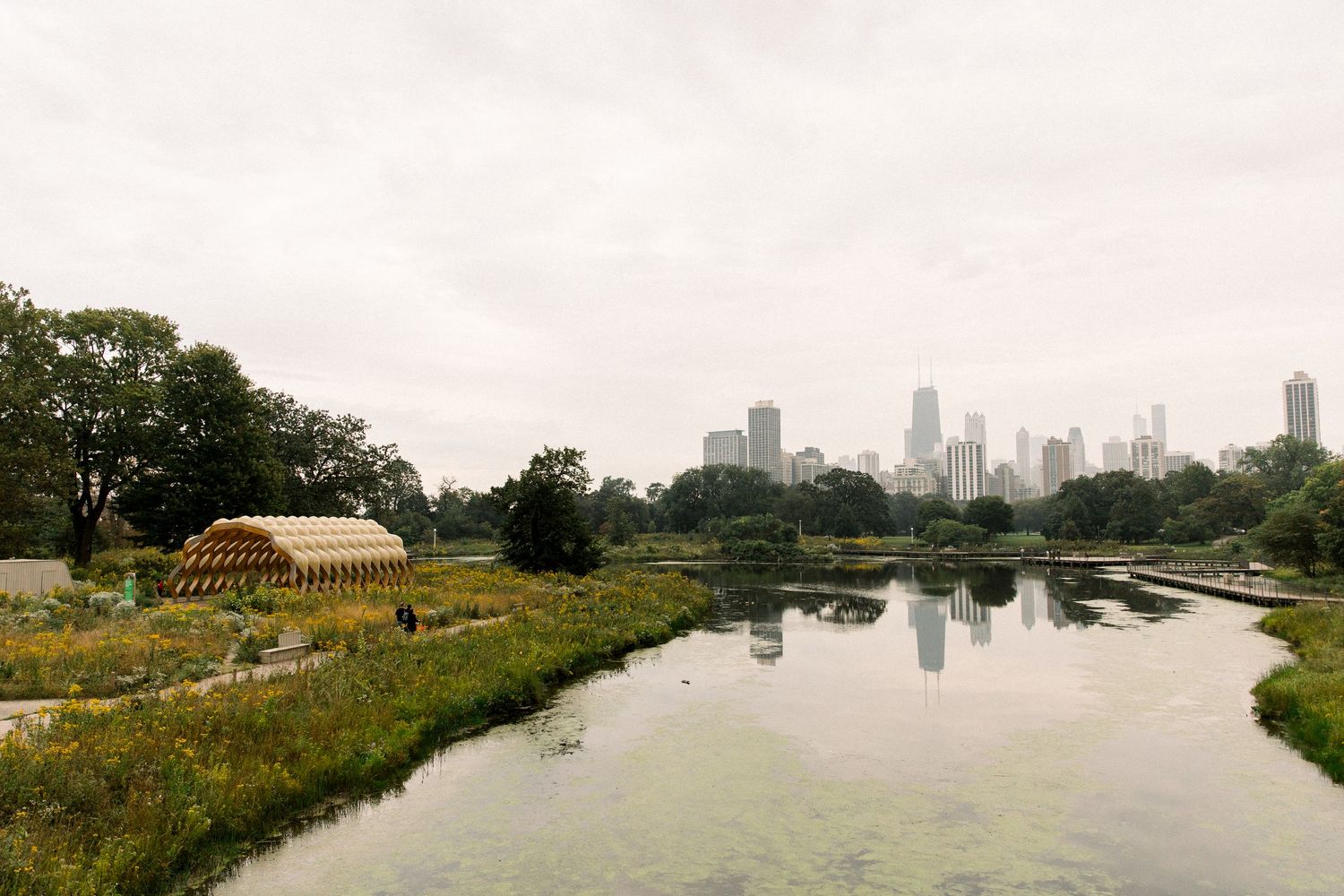 A scenic Chicago park with a yellow geometric pavilion beside a calm river, lush greenery, and city skyline under overcast skies.