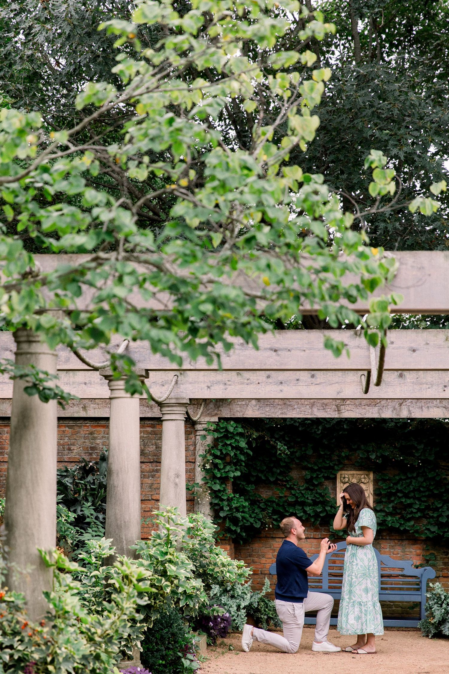 Man proposes on one knee to woman in elegant garden with stone columns and lush greenery.