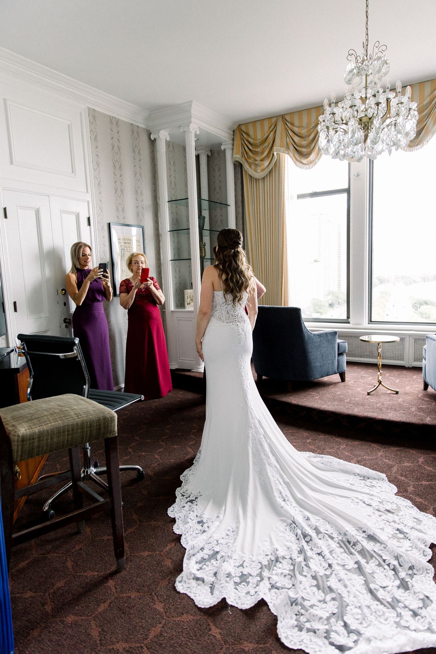 Bride in elegant lace train gown stands by window as bridesmaids in purple and red dresses photograph her in luxury hotel suite.
