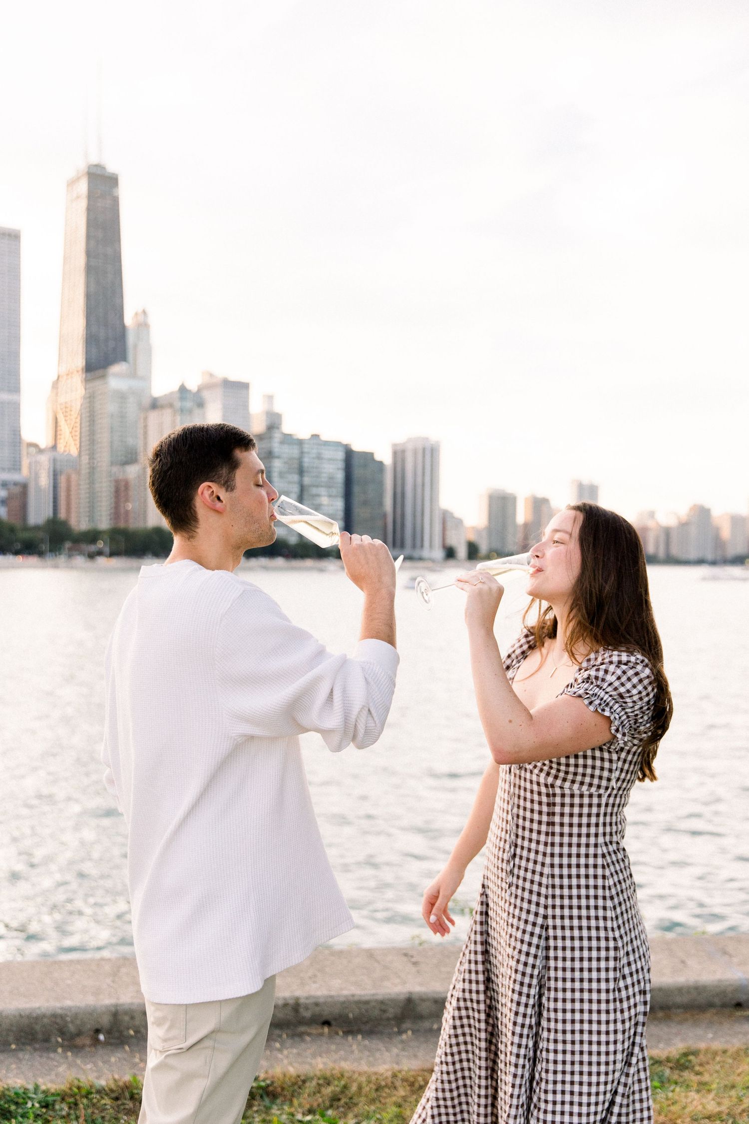 Couple toasting with champagne glasses by Chicago skyline waterfront, man in white shirt, woman in plaid dress.