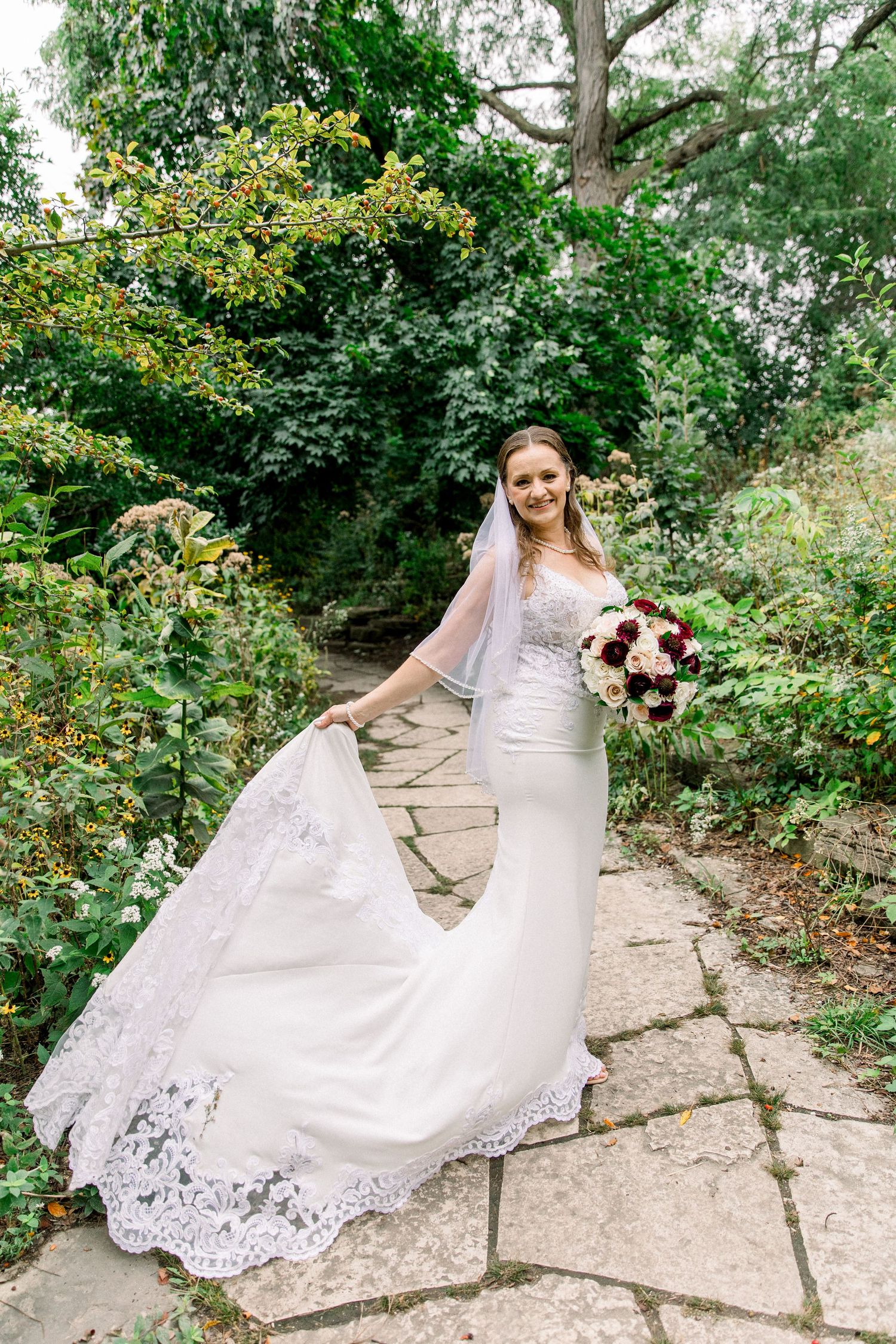 Beautiful bride in lace mermaid wedding dress holding bouquet on stone garden pathway.