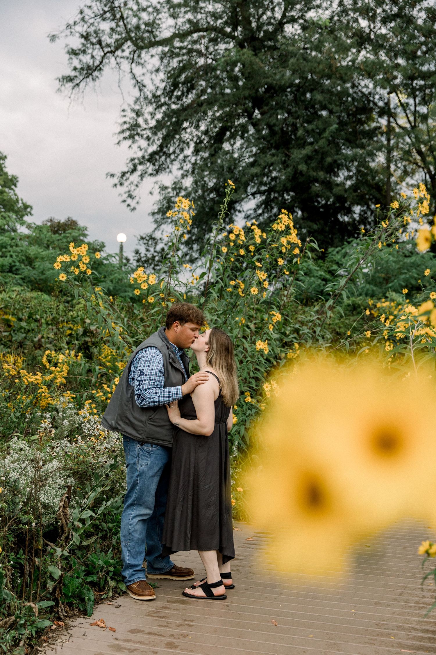 Couple kissing on a wooden path surrounded by vibrant yellow wildflowers and lush green trees in an outdoor garden.